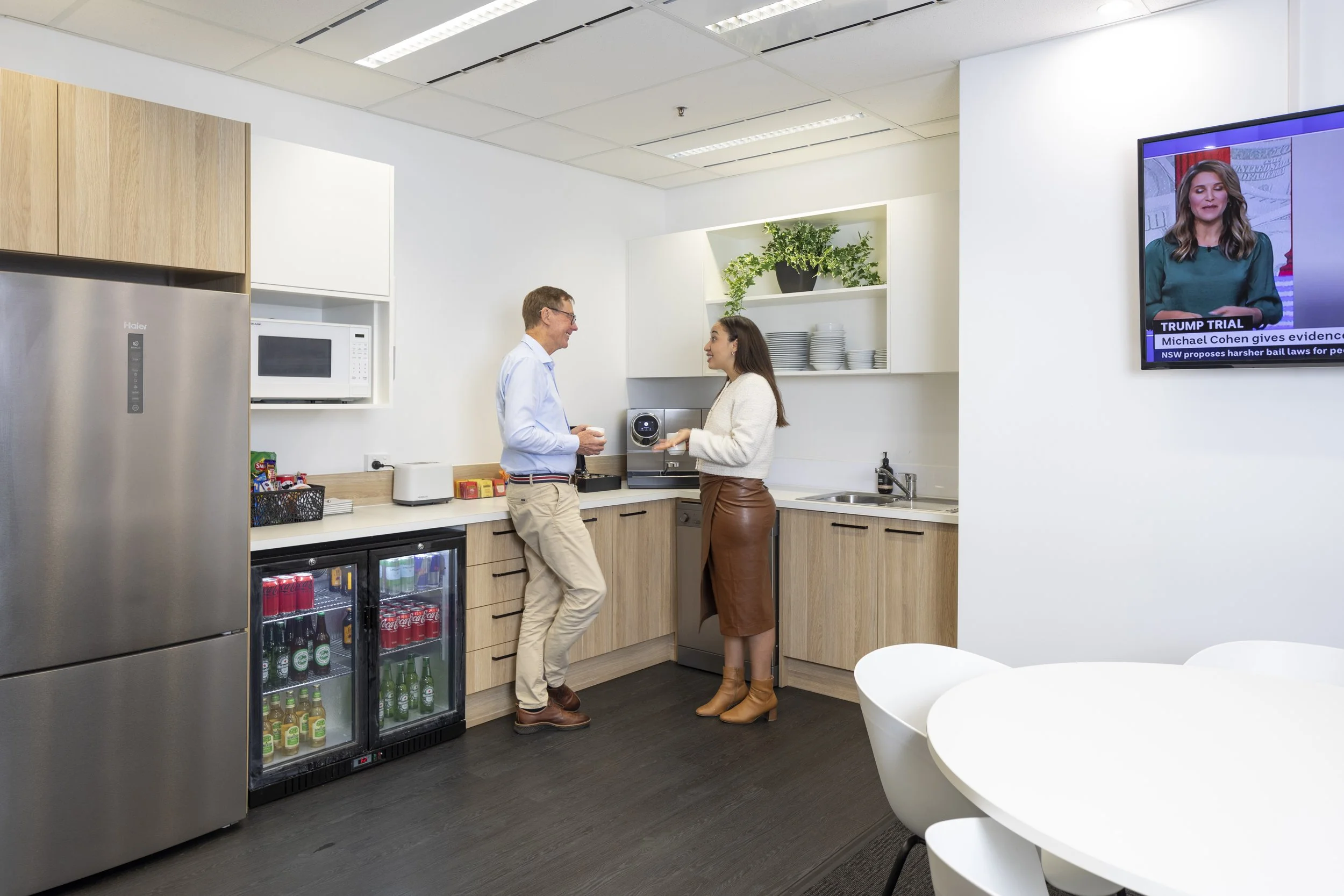 Two people are conversing in an office kitchen. The kitchen has a refrigerator, coffee machine, microwave, snack supplies, lunch table, and a beverage fridge. A wall-mounted TV displays news.