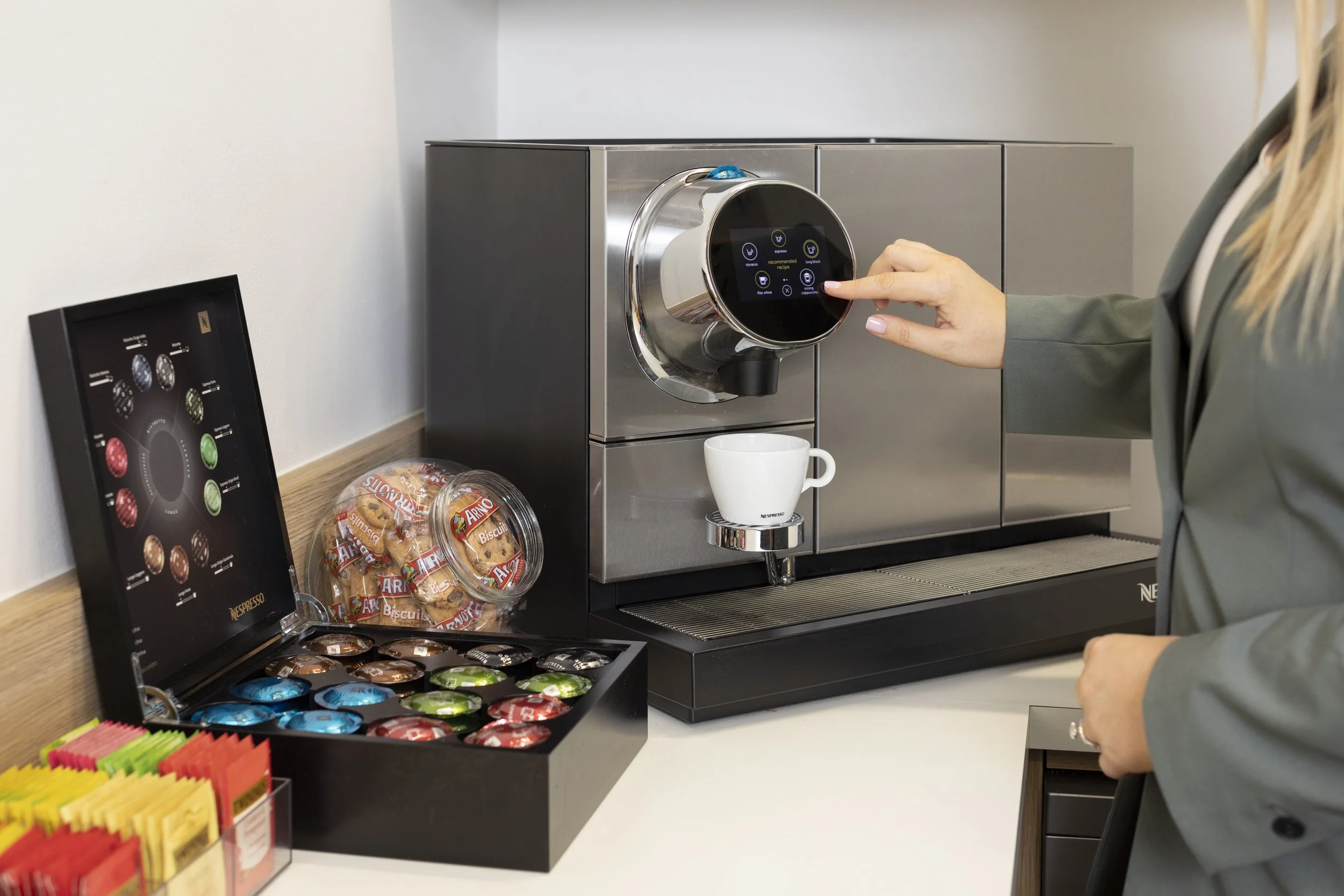 A person presses a button on a modern coffee machine, which is preparing to dispense coffee into a white mug. The coffee machine has a digital display with various options, and there are packets of biscuits and a selection of coffee capsules nearby on the counter.