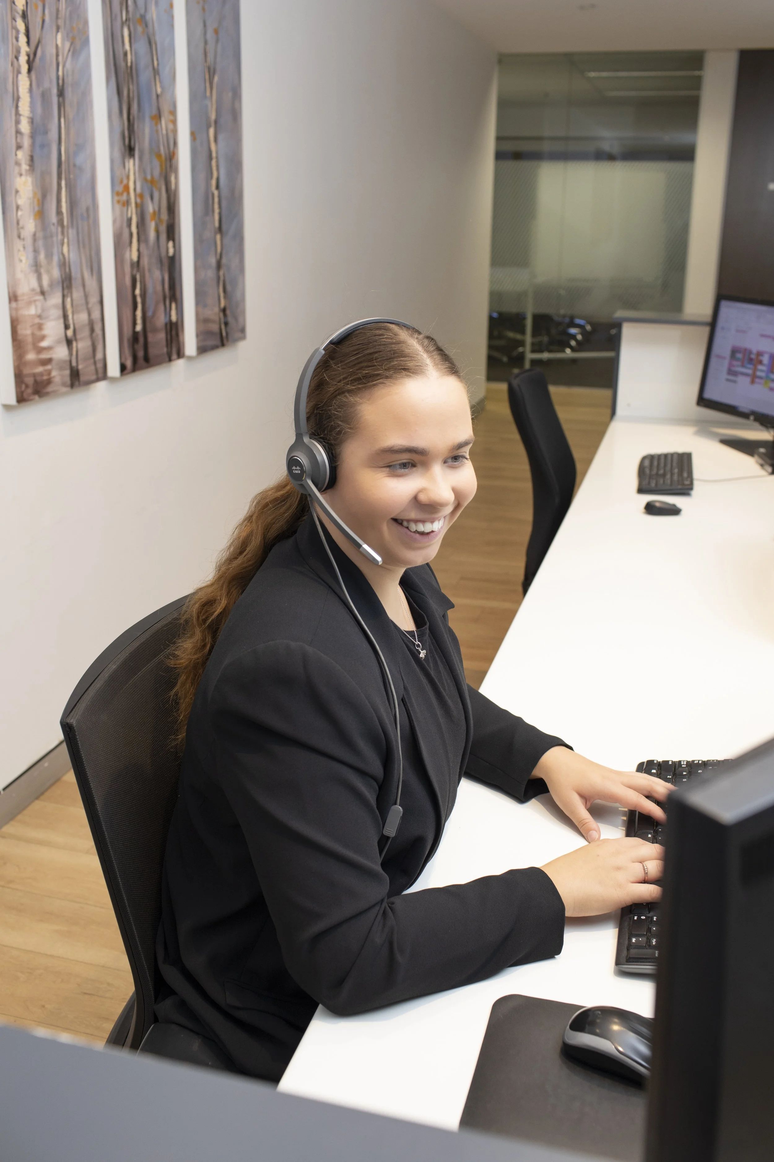 A woman with long brown hair smiling while working at a computer in an office. She wears a black blazer and a headset, and is seated at a white desk with a keyboard and mouse.