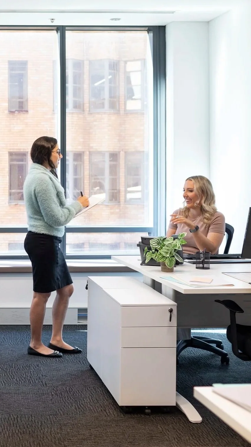 Two women conversing in an office, one standing with a notepad and the other sitting at a desk smiling, with a large window and cityscape background.