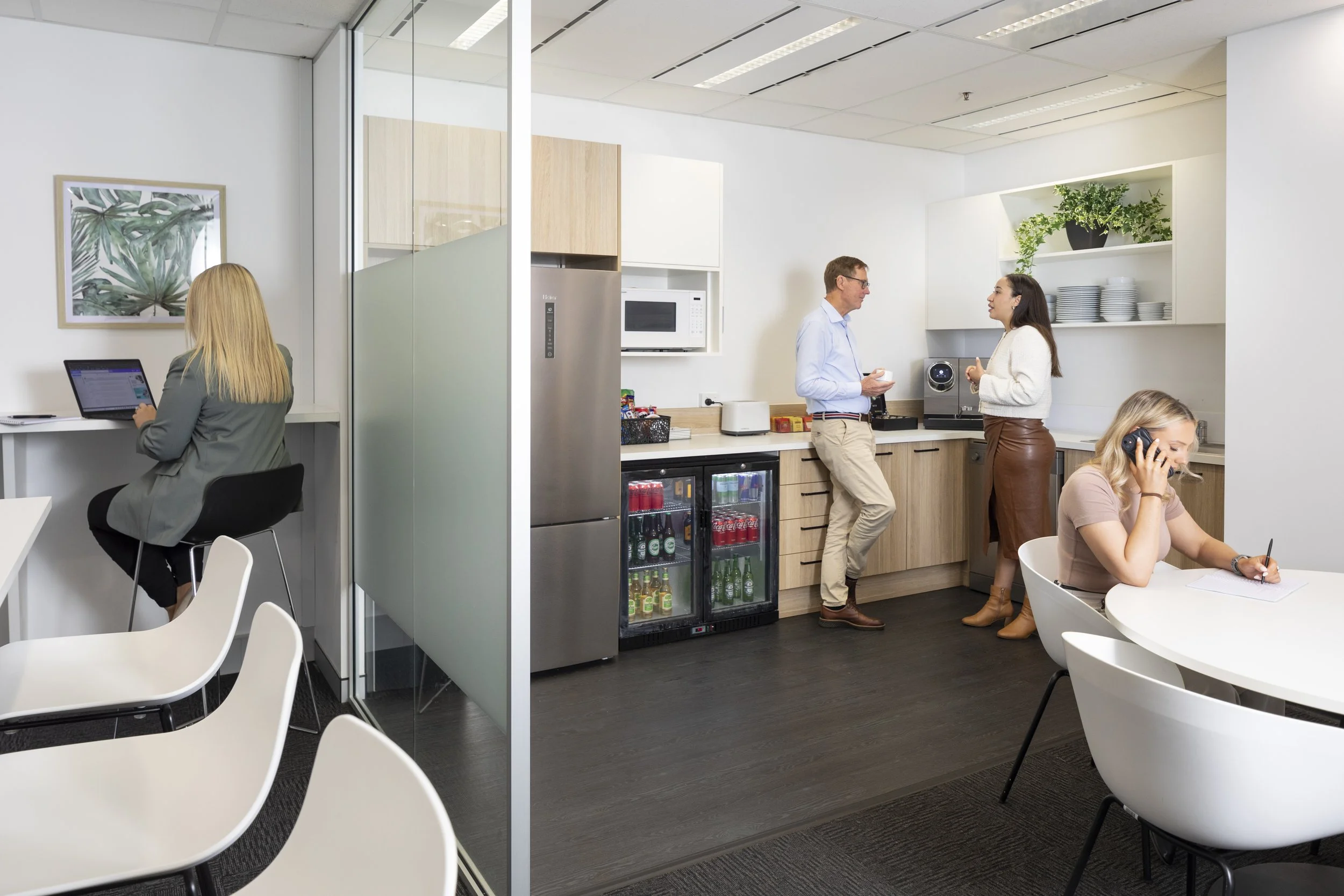 Office kitchen area with three people, a woman working on her laptop, a man and woman talking near the counter, and a woman sitting and talking on the phone, with shelves, plates, and a beverage fridge in the background.