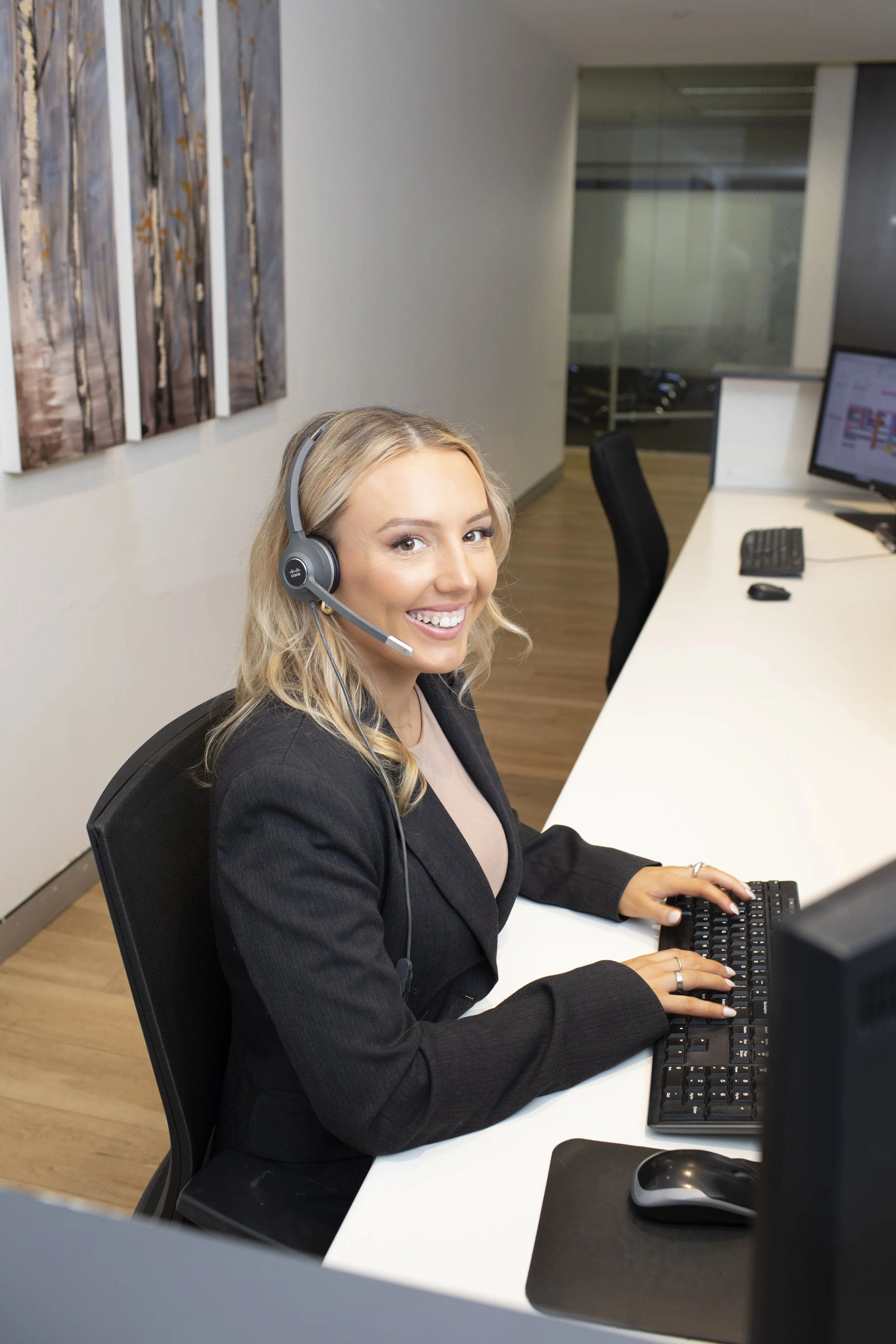 Smiling woman with long blonde hair wearing a headset, using a keyboard and mouse at an office desk.