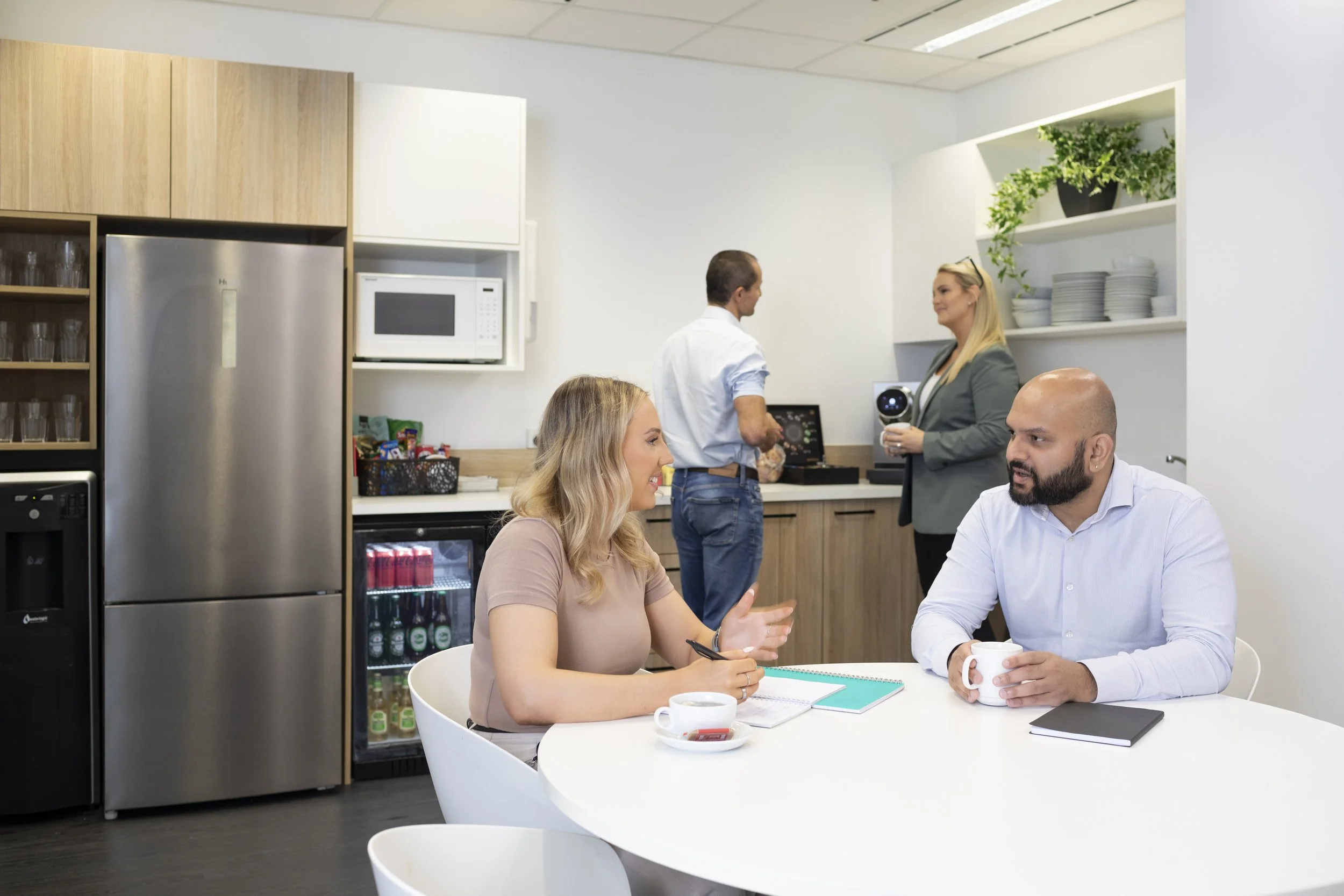 People in a modern office kitchen and meeting room, engaging in conversation. A woman and a man are seated at a white table with cups and notebooks, while two others are in the background near the kitchen counter and cabinets.