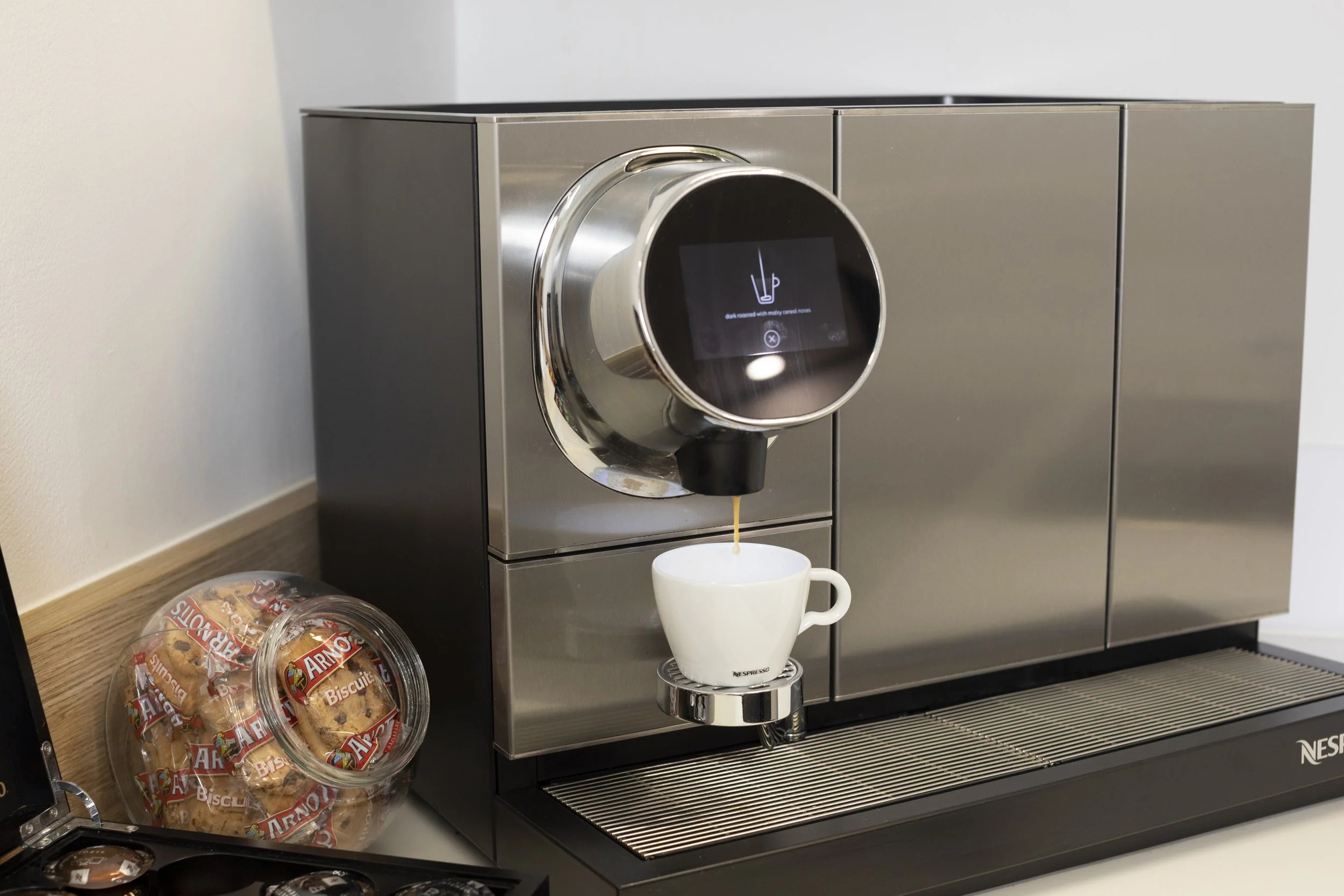 A modern coffee machine dispensing coffee into a white mug, with a container of Arnotts Biscuits on a wooden surface to the side.