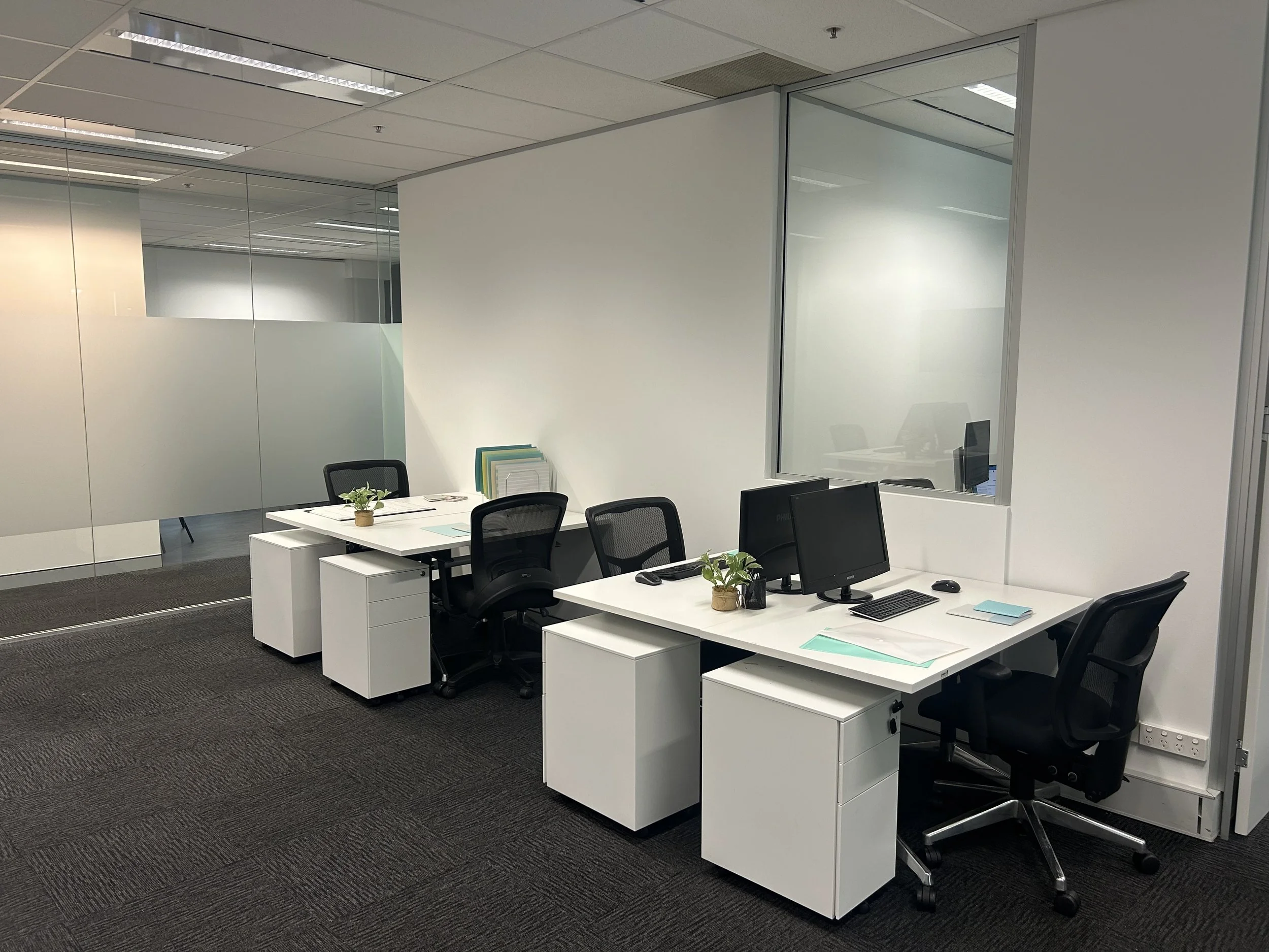 An office with white desks, black chairs, desktop computers, and a small plant. A glass partition wall with a white wall behind it, and ceiling lights.