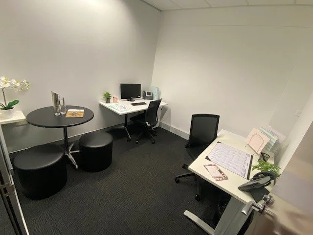 Small office space with two desks, one with a computer and the other with documents, chairs, and potted plants, in a minimalist setting.