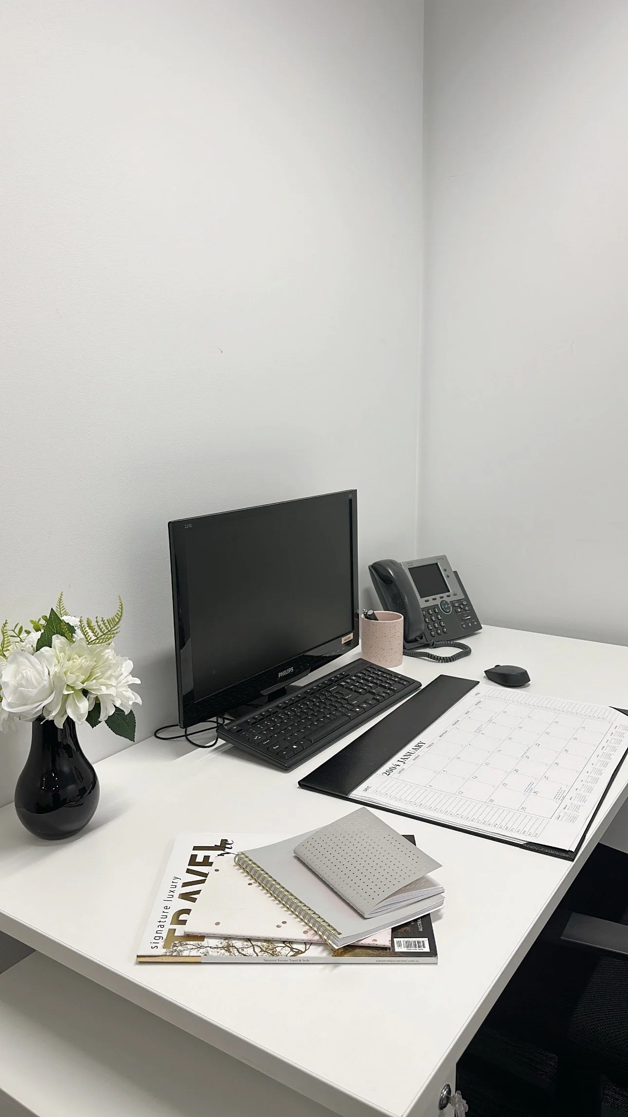 Office desk with a computer monitor, keyboard, mouse, desk phone, planner, notebooks, and a vase with white flowers against a white wall.