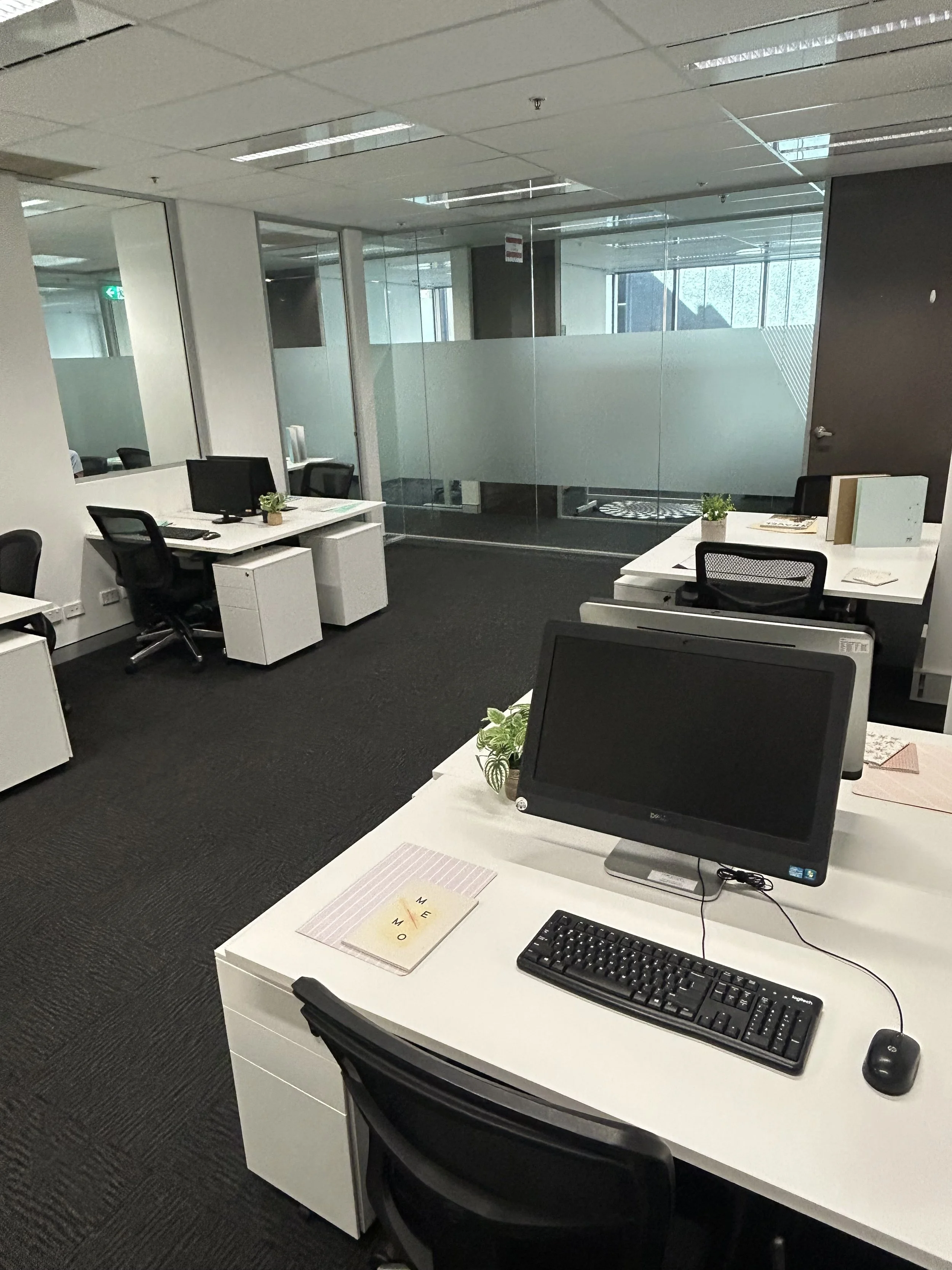 An office with multiple white desks, black chairs, desktop computers, and some potted plants. Glass walls separate different areas in the office.