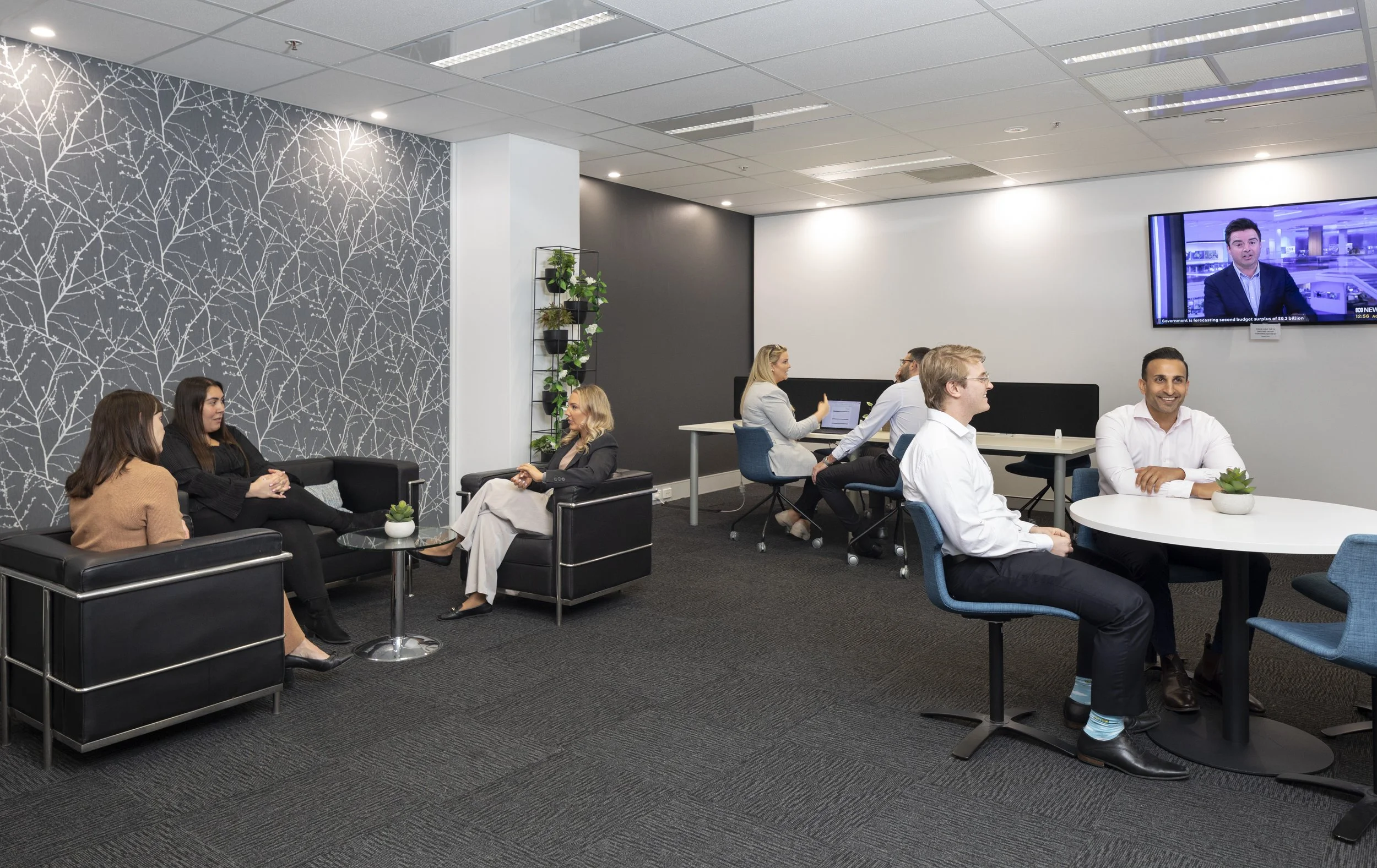A modern office waiting or lounge area with five women and two men, some sitting on black leather sofas and others at a round table, engaged in conversation or working on laptops. There is a wall with white branch pattern wallpaper, a plant stand with potted plants, and a wall-mounted television displaying a news anchor.