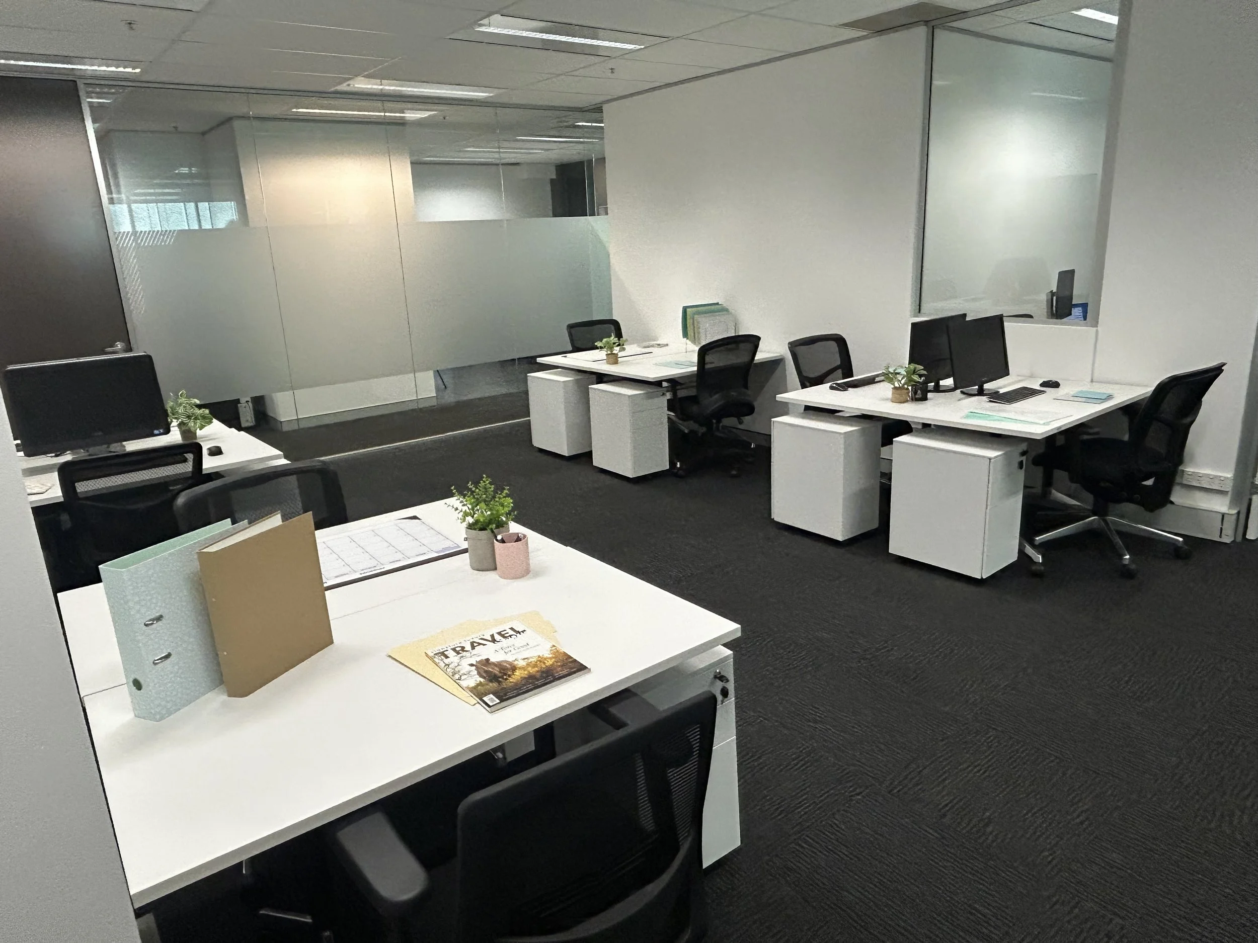 An office space with white desks, black chairs, several computer monitors, office supplies, small potted plants, and glass partitions.