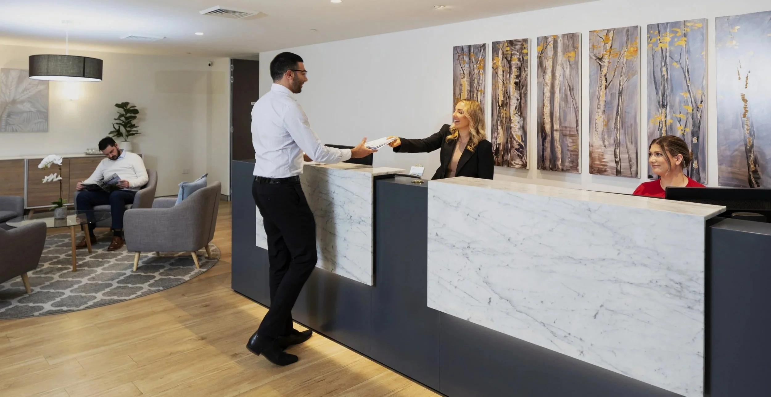 Reception area with a man handing paperwork to a receptionist. Two women are seated nearby, one reading and one smiling. The decor includes modern paintings, a potted plant, and cozy seating.