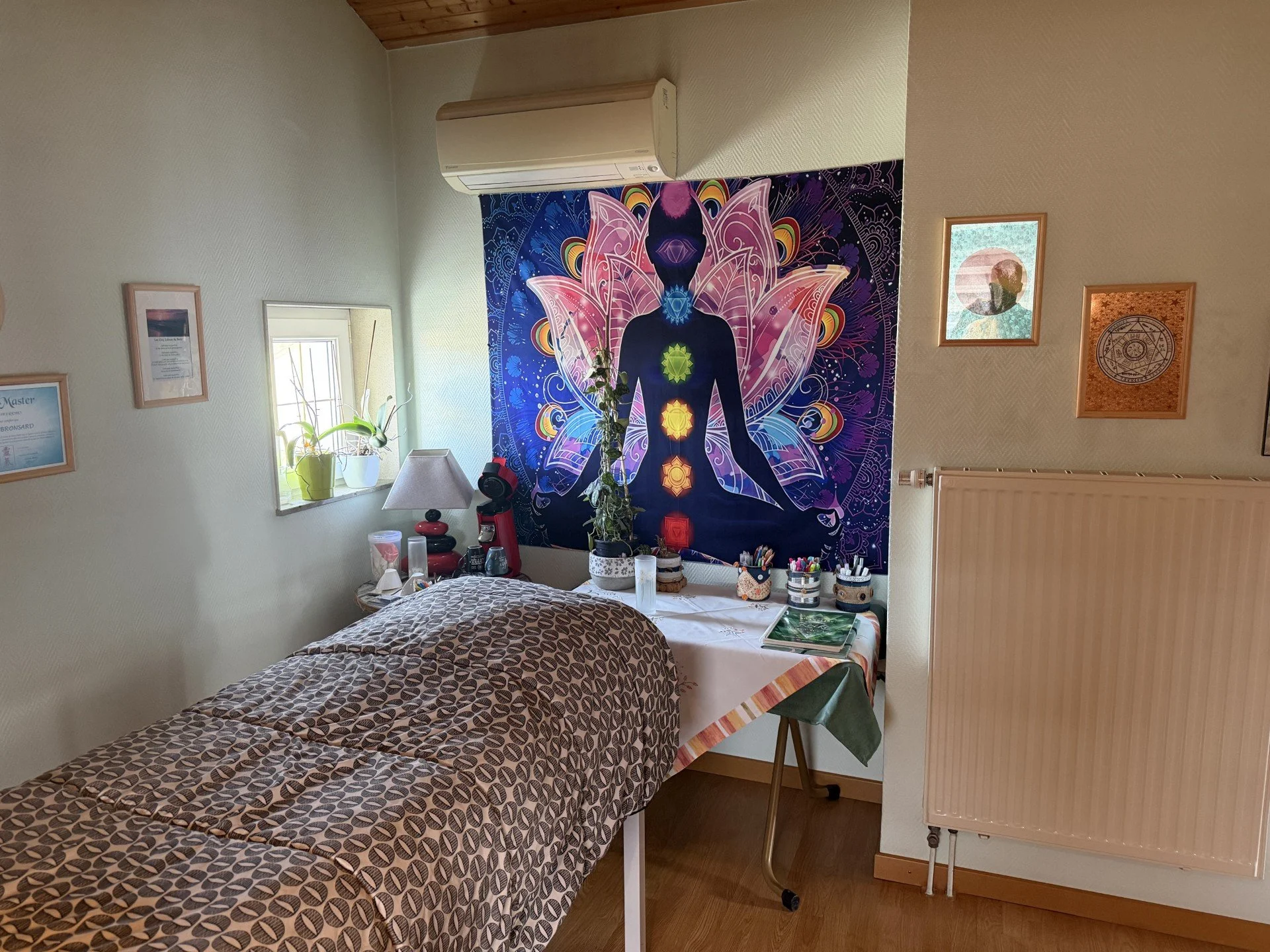 Chambre avec un lit recouvert d'une couverture à motifs ovals, un mur décoré d'une grande tenture de yoga avec un corps humain silhouette et chakras, une plante en pot sur la fenêtre, un lampadaire, un radiateur et quelques cadres suspendus au mur.