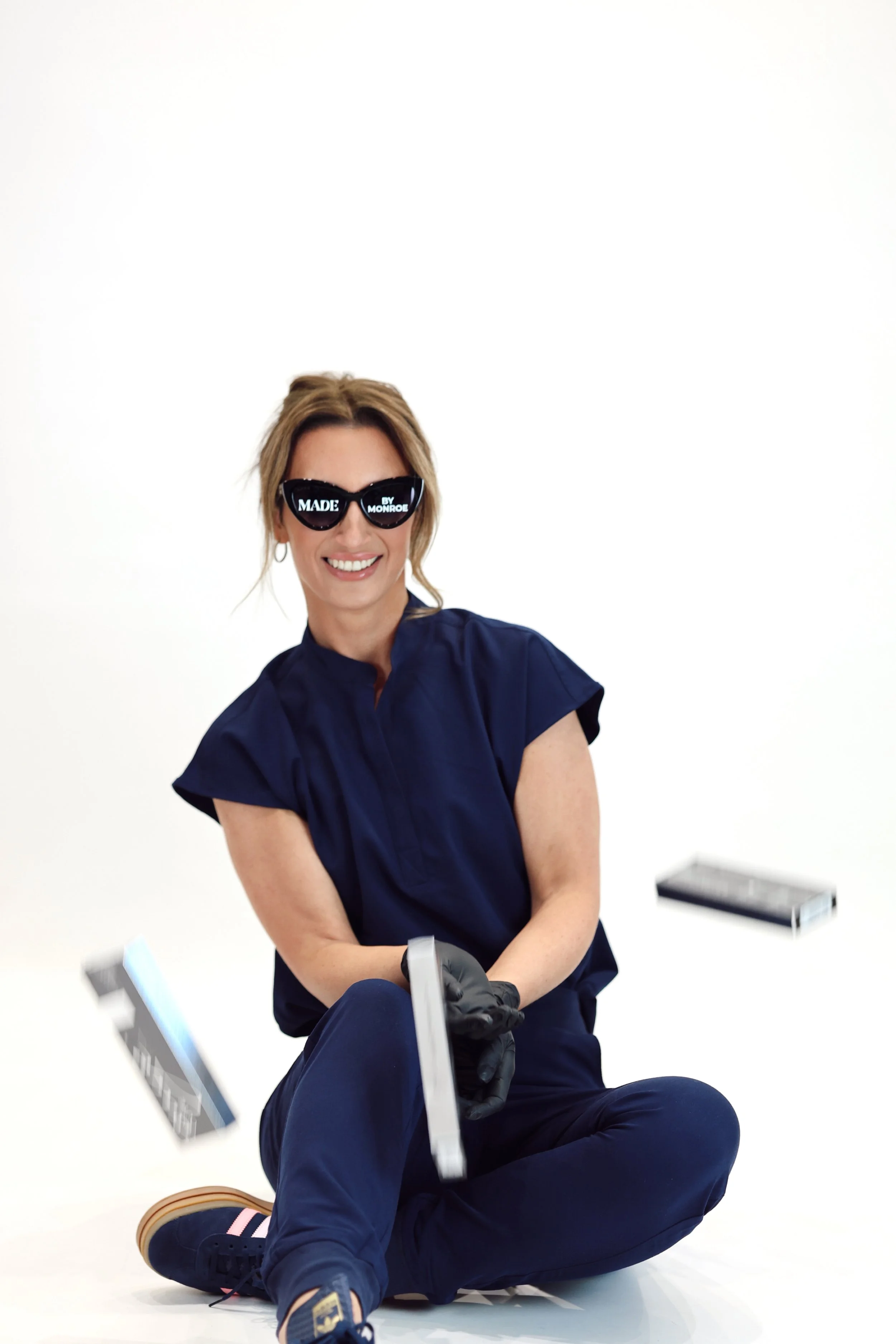 Smiling woman wearing sunglasses and a navy blue outfit, sitting on the floor, surrounded by flying papers, white background.