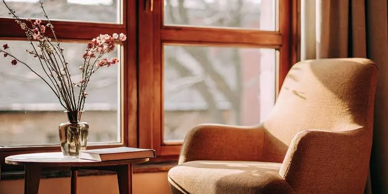 A cozy corner with a beige armchair next to a wooden table with a vase of pink flowers, in front of a window with wooden trim showing blurred trees outside.