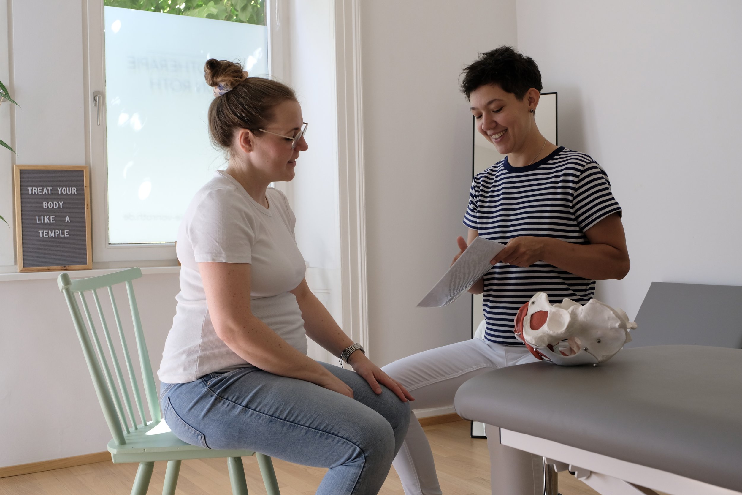 Zwei Frauen in einem medizinischen Raum, eine sitzt auf einem Stuhl und die andere hält eine Broschüre. Im Hintergrund ist ein Fenster mit einem Schild, das auf Englisch steht, und ein Modell eines menschlichen Beckens auf dem Tisch.