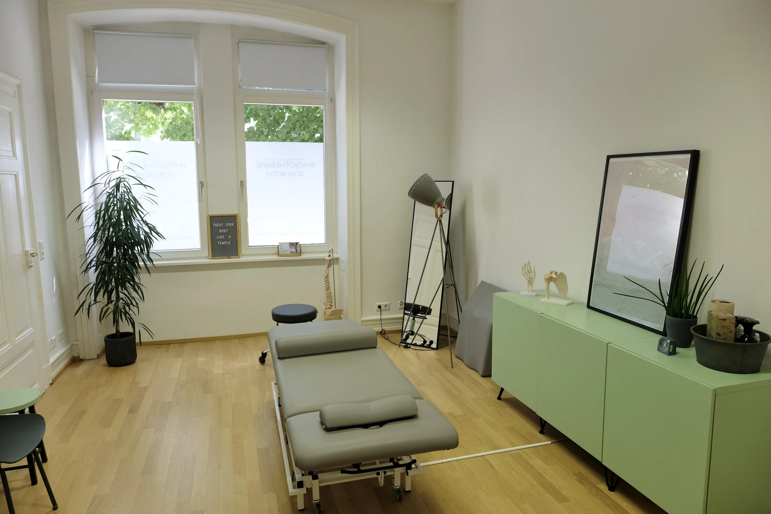 Behandlungsraum mit Untersuchungsliege, Pflanzen, Spiegel, Stehleuchte, Fenster mit grünem Ausblick, Dekorationsgegenständen auf einer grünen Schrank.