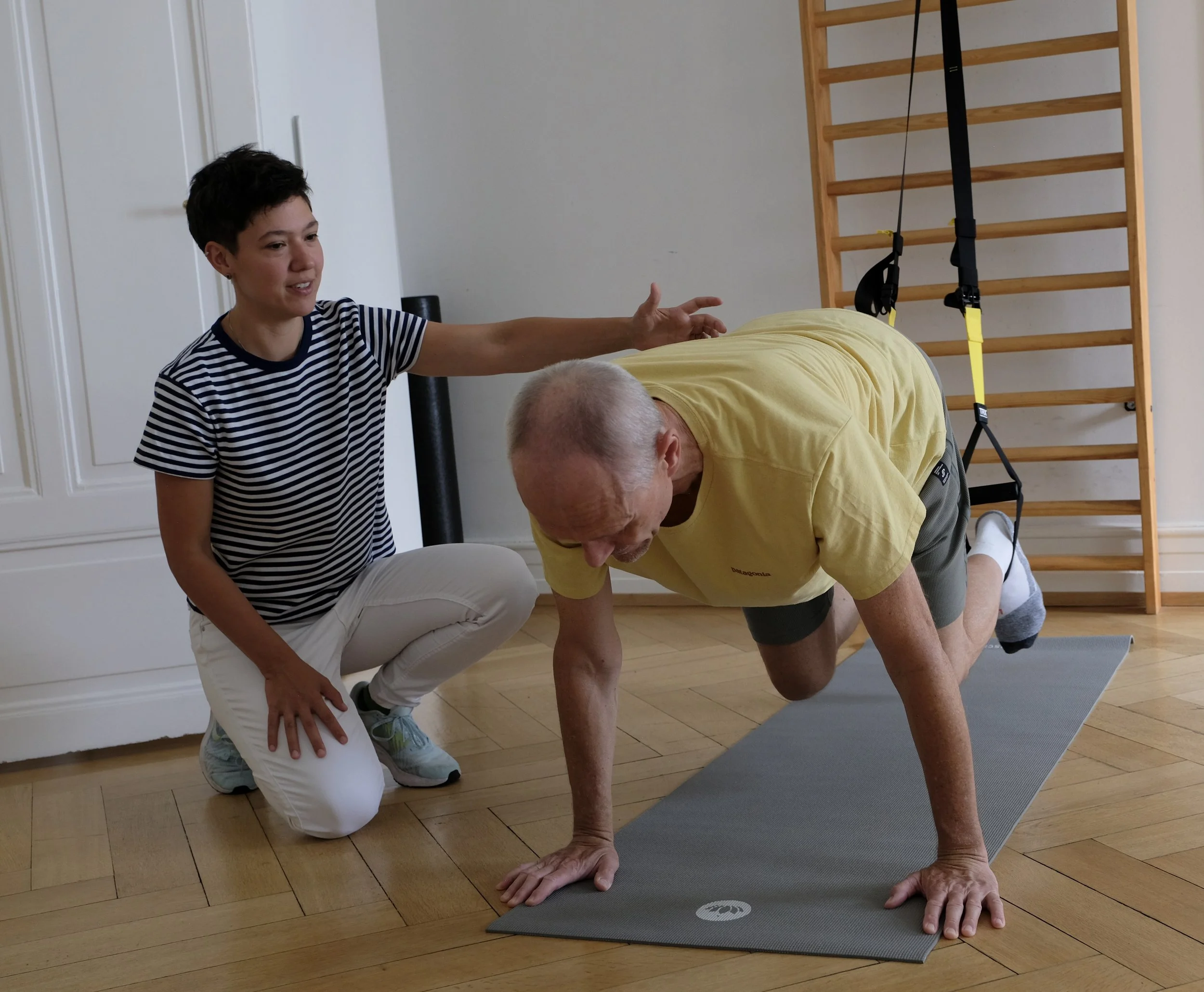 Eine junge Frau hilft einem alten Mann bei einer Yoga-Übung in einem Fitnessraum, während sie eine Hand auf seinen Rücken legt. Der Mann befindet sich in einer Plank-Position auf einer Yogamatte, mit einem ausgestreckten Arm und einem gestreckten Bei