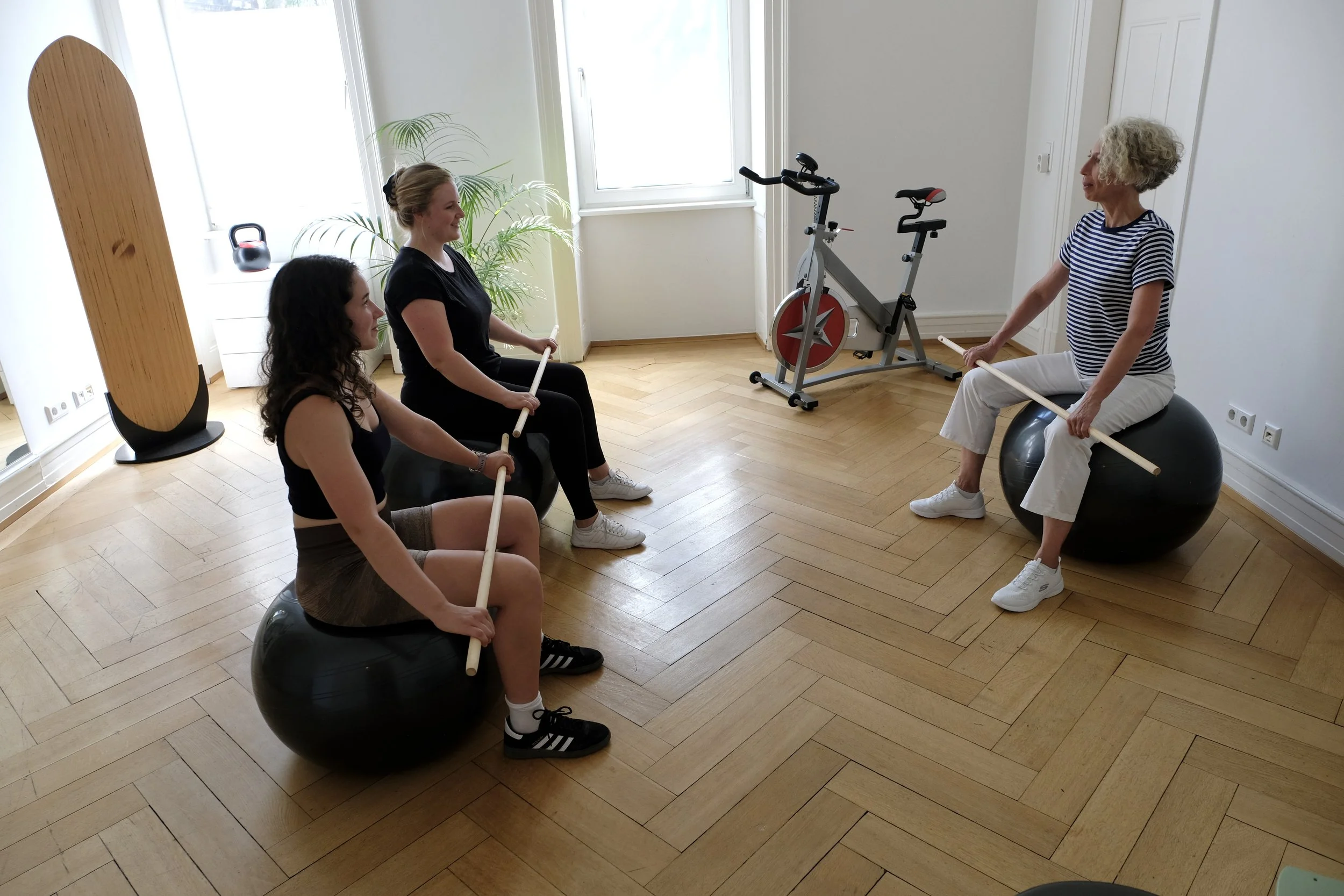 Eine Gruppe von vier Frauen sitzt auf Gymnastikbällen in einem Raum, während eine ältere Frau auf einem Gymnastikball sitzt und ihnen mit einem Holzstab zeigt. Im Hintergrund befinden sich ein Fahrrad-Ergometer, eine Pflanze und große Fenster.
