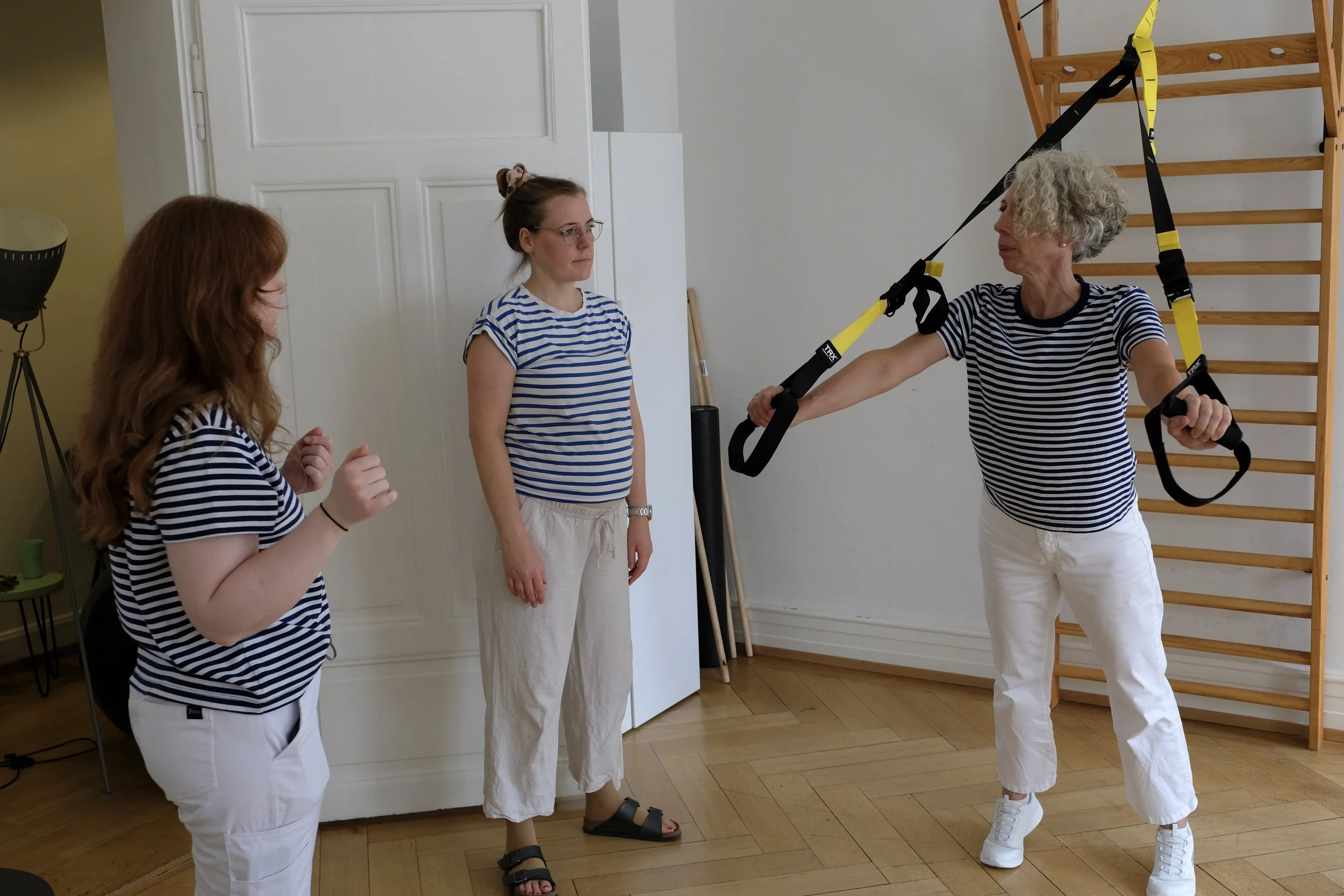 Drei Frauen in einem Raum mit weißen Wänden und Holzfußboden, die eine Trainingseinheit mit Gymlandschafts-Equipment durchführen. Zwei Frauen tragen gestreifte Shirts, eine trägt weiße Hosen, die andere schwarze Sandalen. Eine Frau benutzt TRX-Schlin