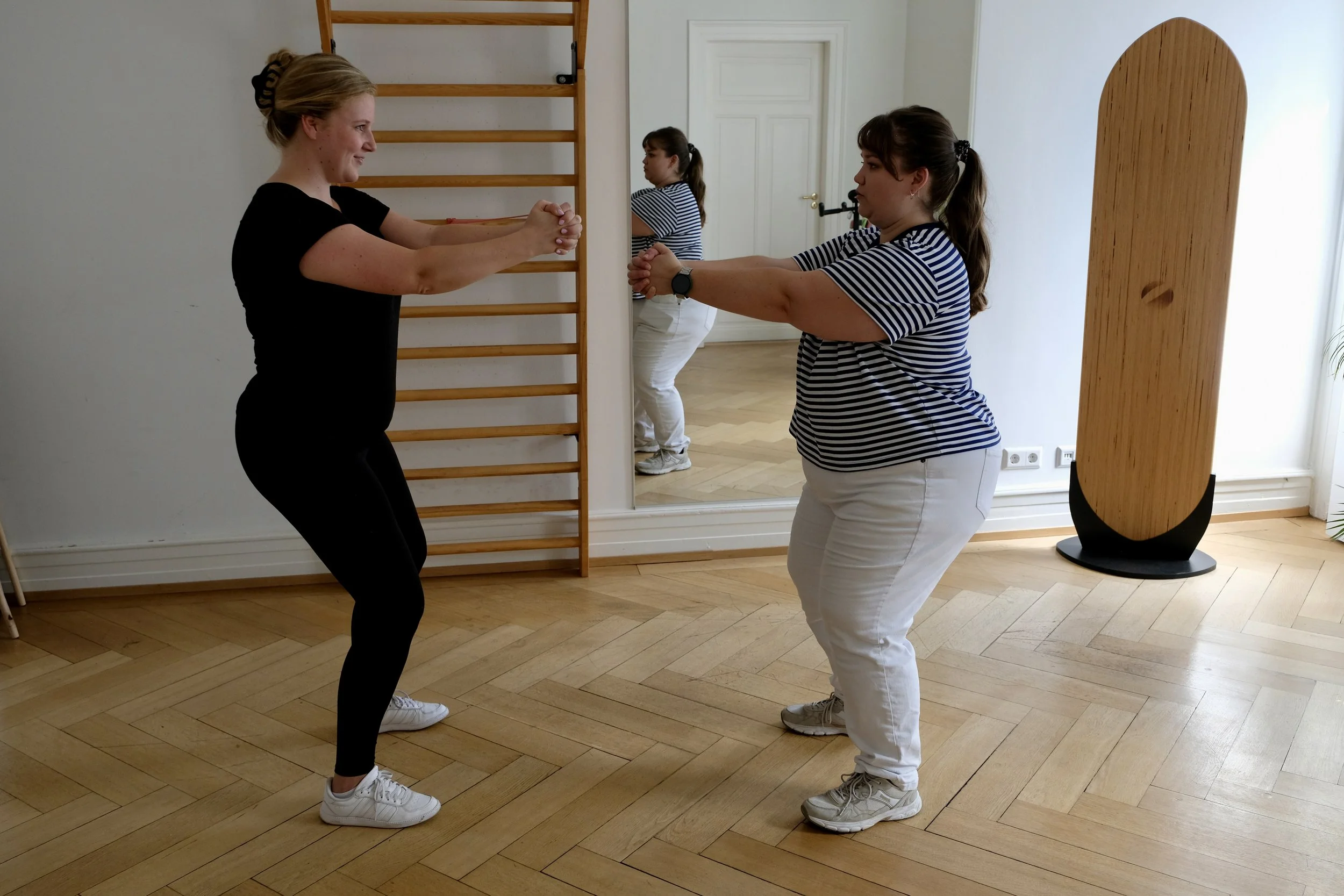 Zwei Frauen trainieren im Fitnessraum, stehen sich mit angewinkelten Armen gegenüber und halten sich an den Händen, während eine dritte Frau im Hintergrund ebenfalls übt.