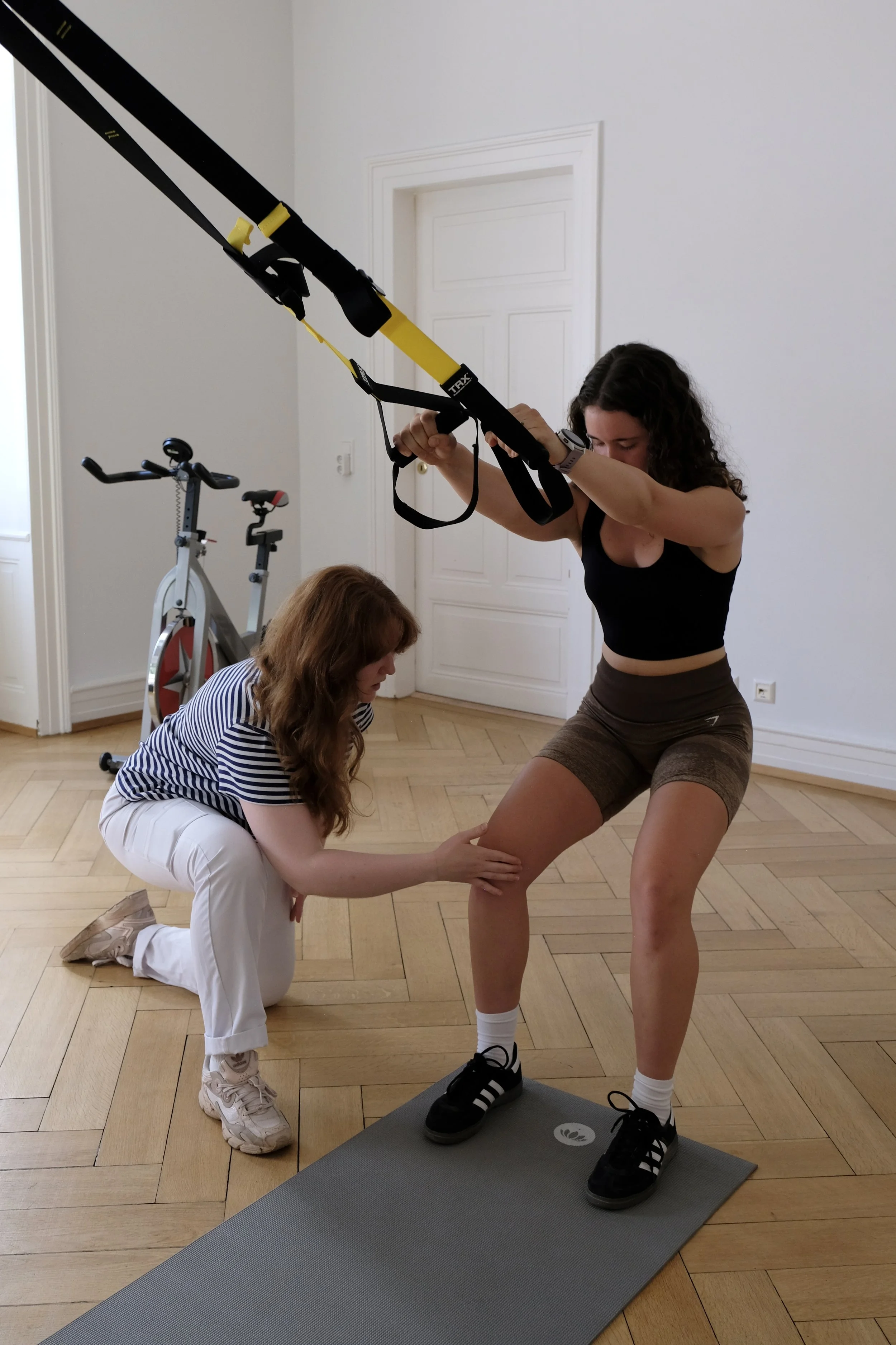 Eine junge Frau macht eine Balanceübung auf einem Balance-Board, unterstützt von einer Trainerin, die ihr die Knie überprüft, während eine zweite assistierende Person im Hintergrund eine Maschine im Fitnessraum benutzt.