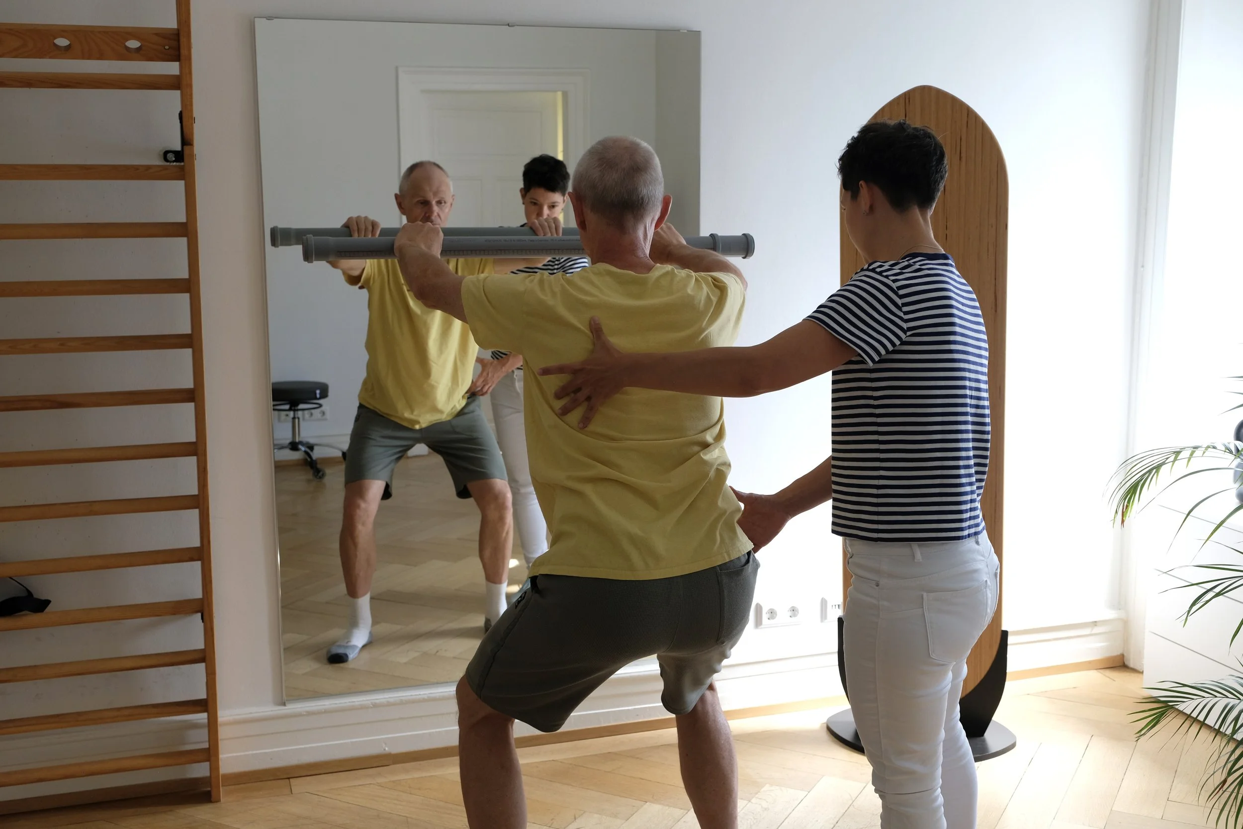 Ein Personal Trainer hilft einem älteren Mann beim Gleichgewichtstraining vor einem Spiegel, während eine junge Frau assistiert.
