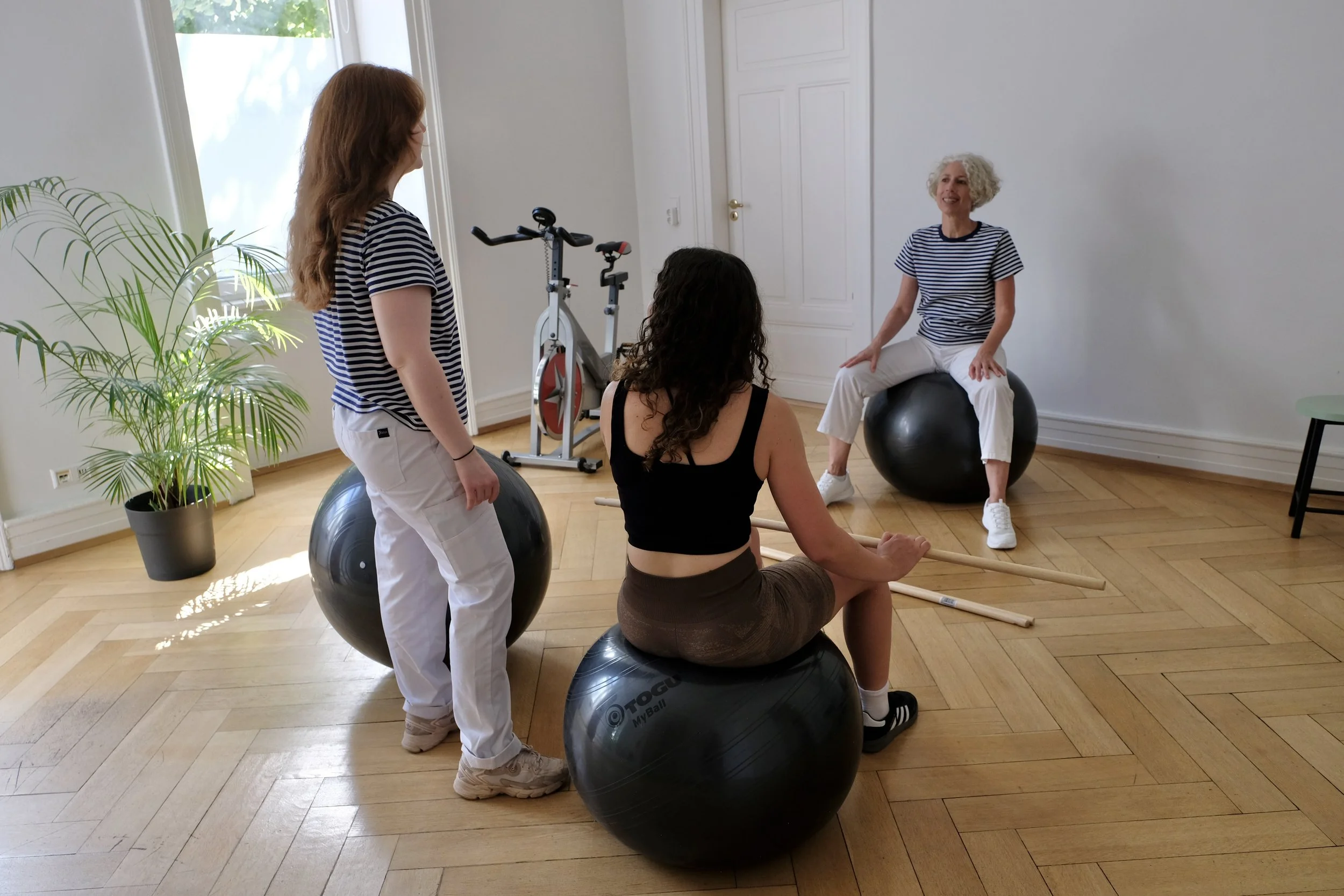 Fünf Frauen in einem Raum, der für eine Fitness- oder Physiotherapie-Sitzung eingerichtet ist, mit Gymnastikbällen, einem Fahrrad und Spielholzstäben. Eine Frau sitzt auf einem Gymnastikball, während die anderen um sie herum stehen oder sitzen.