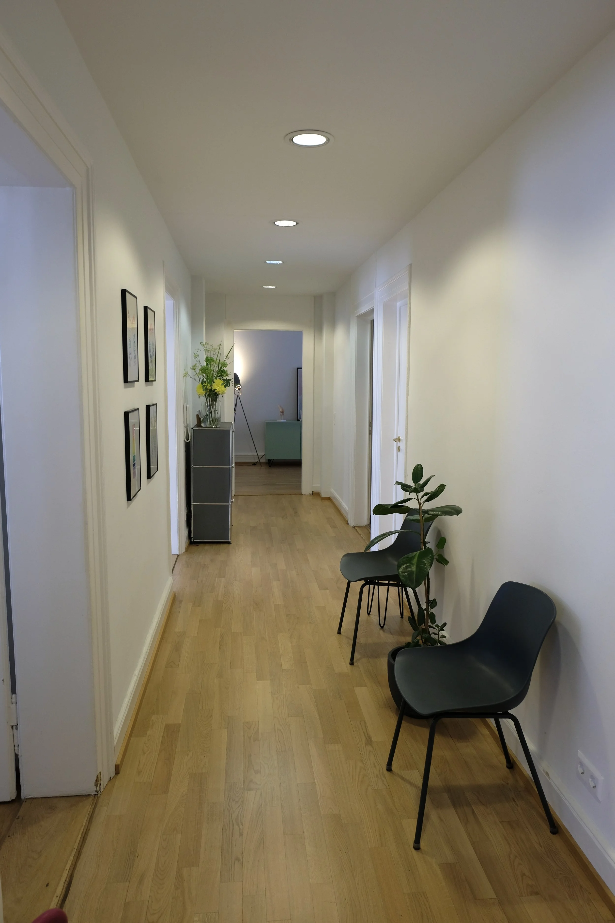 Wartebereich: Ein heller Flur mit Stühlen, einer Pflanze und Bildern an der Wand, im Hintergrund ein Tisch und weitere Einrichtung.