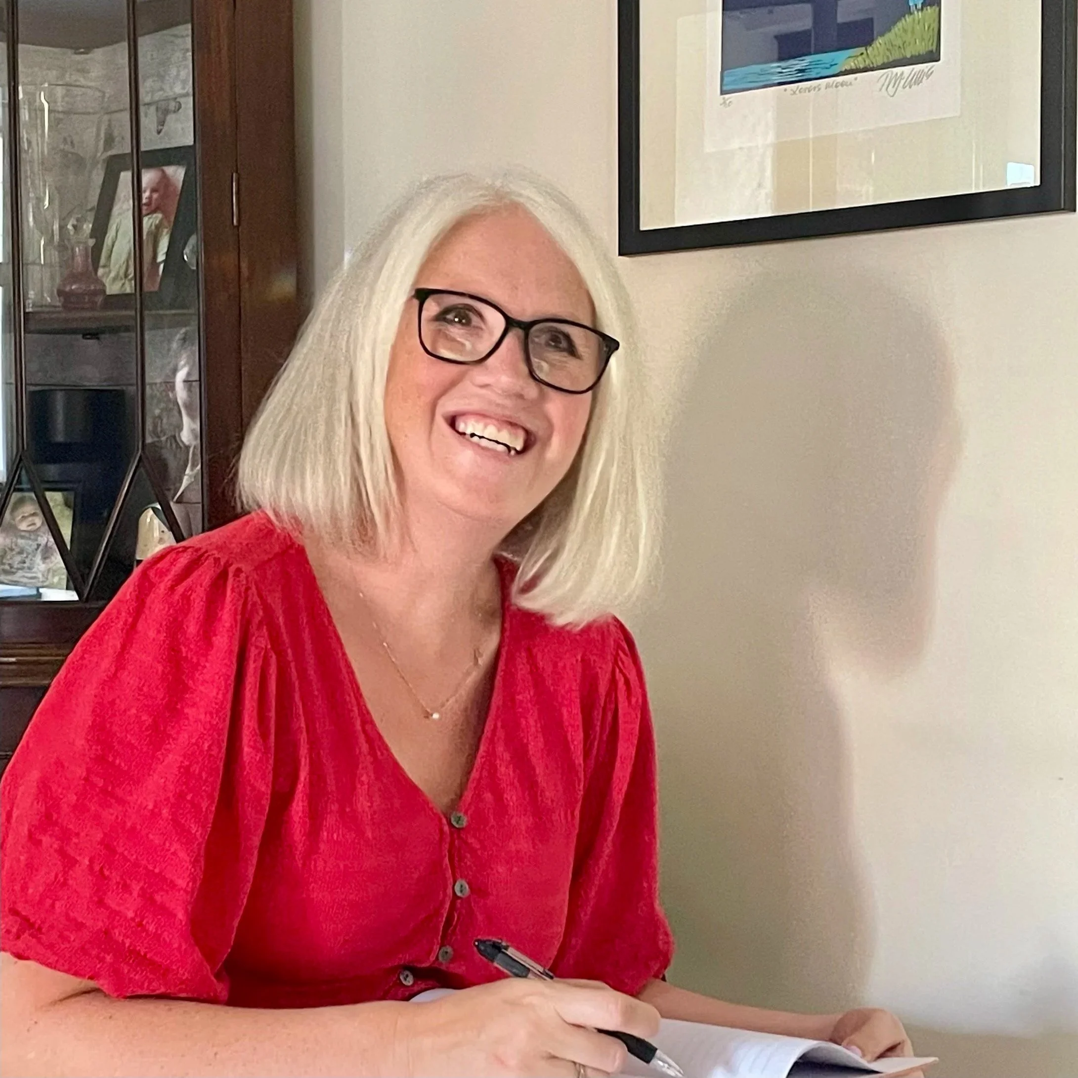 Close-up of smiling white-haired woman wearing glasses and red blouse, holding a pen and writing on a paper with framed artwork and a glass display cabinet behind her.