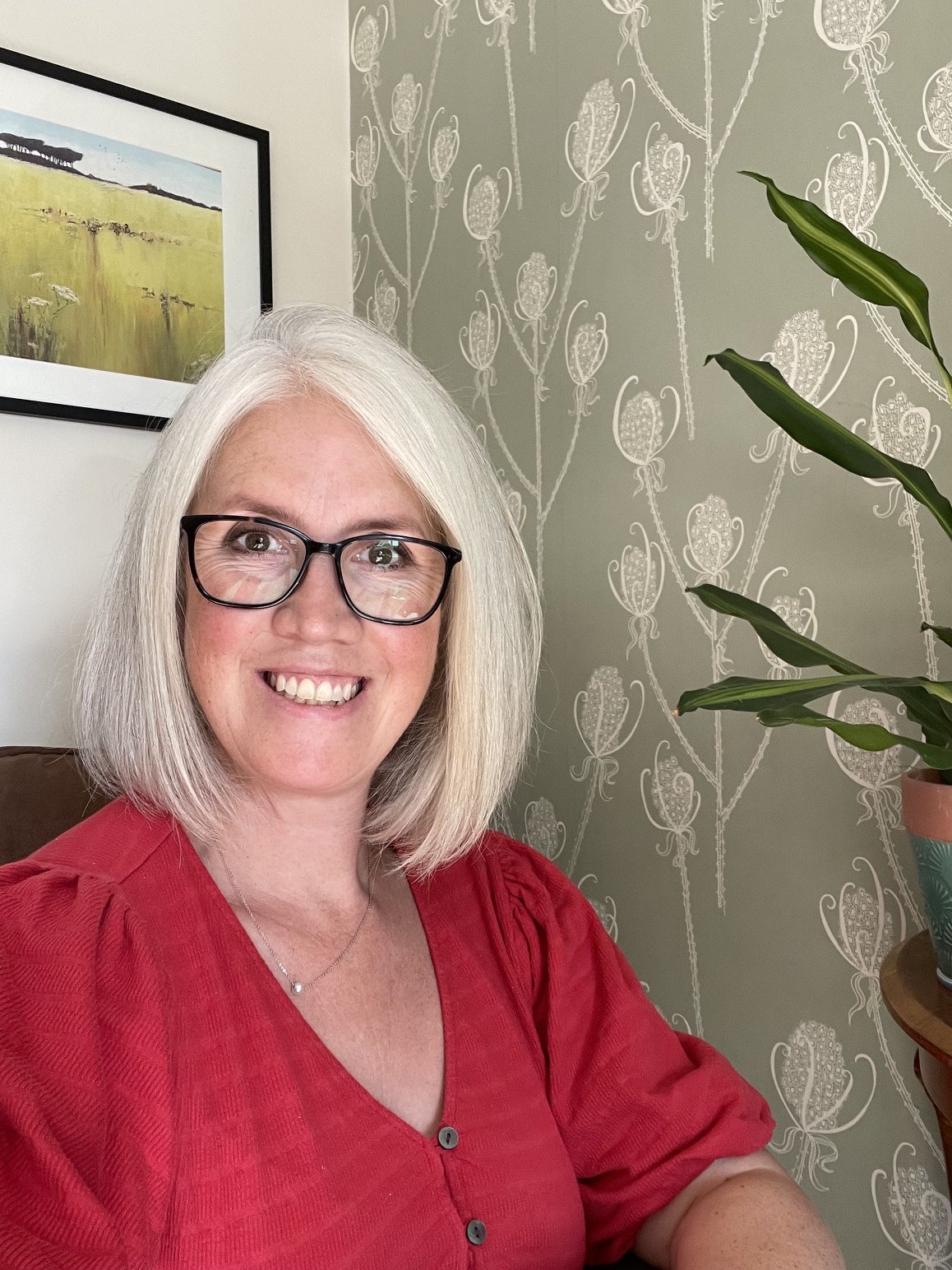 A woman with white hair and glasses smiling, wearing a red top, sitting near a potted plant, with framed landscape artwork and patterned wallpaper in the background.