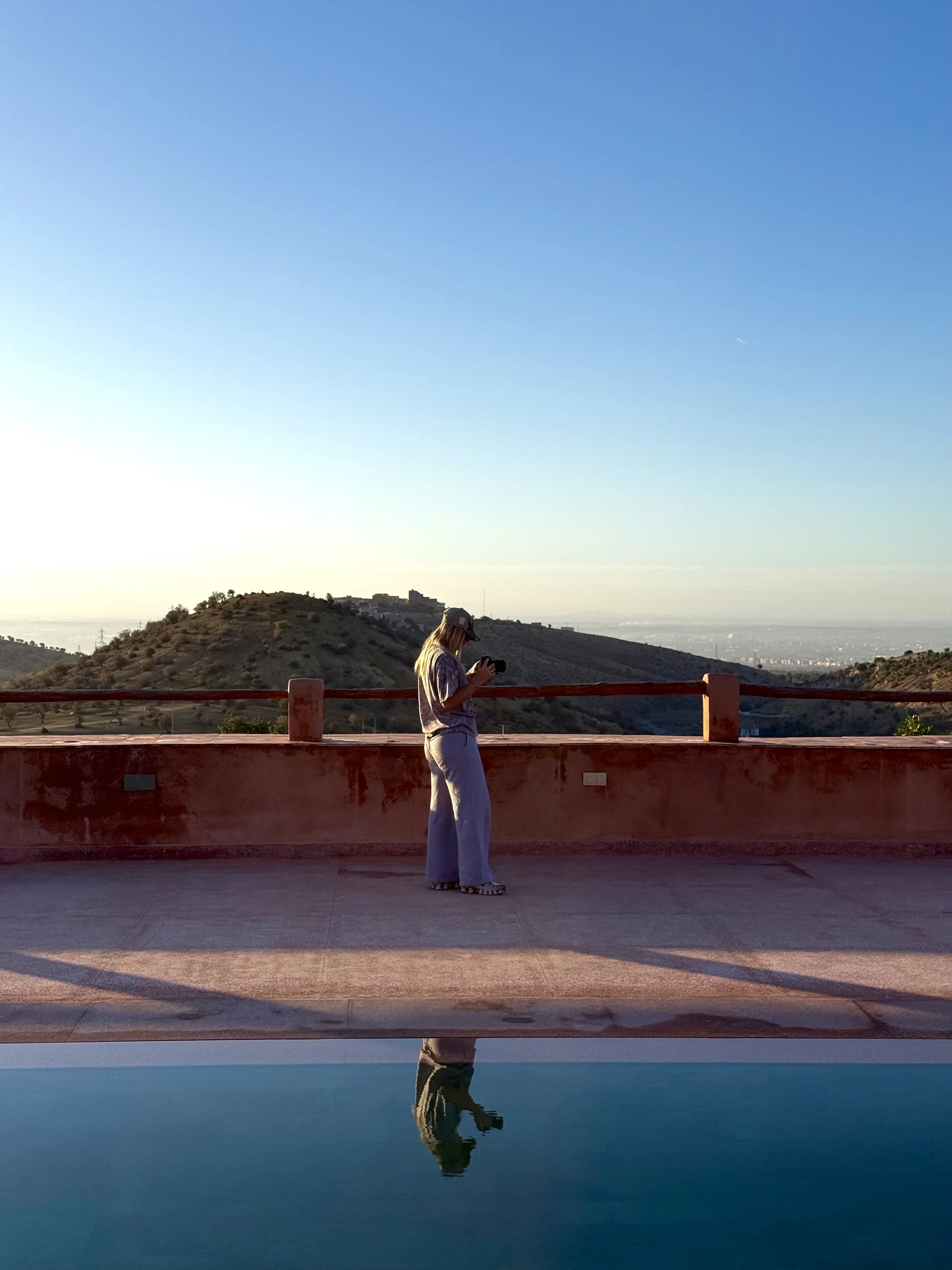 A woman standing on a deck, looking at her phone, with a landscape of hills and a distant house under a clear blue sky, reflection in a pool of water below.