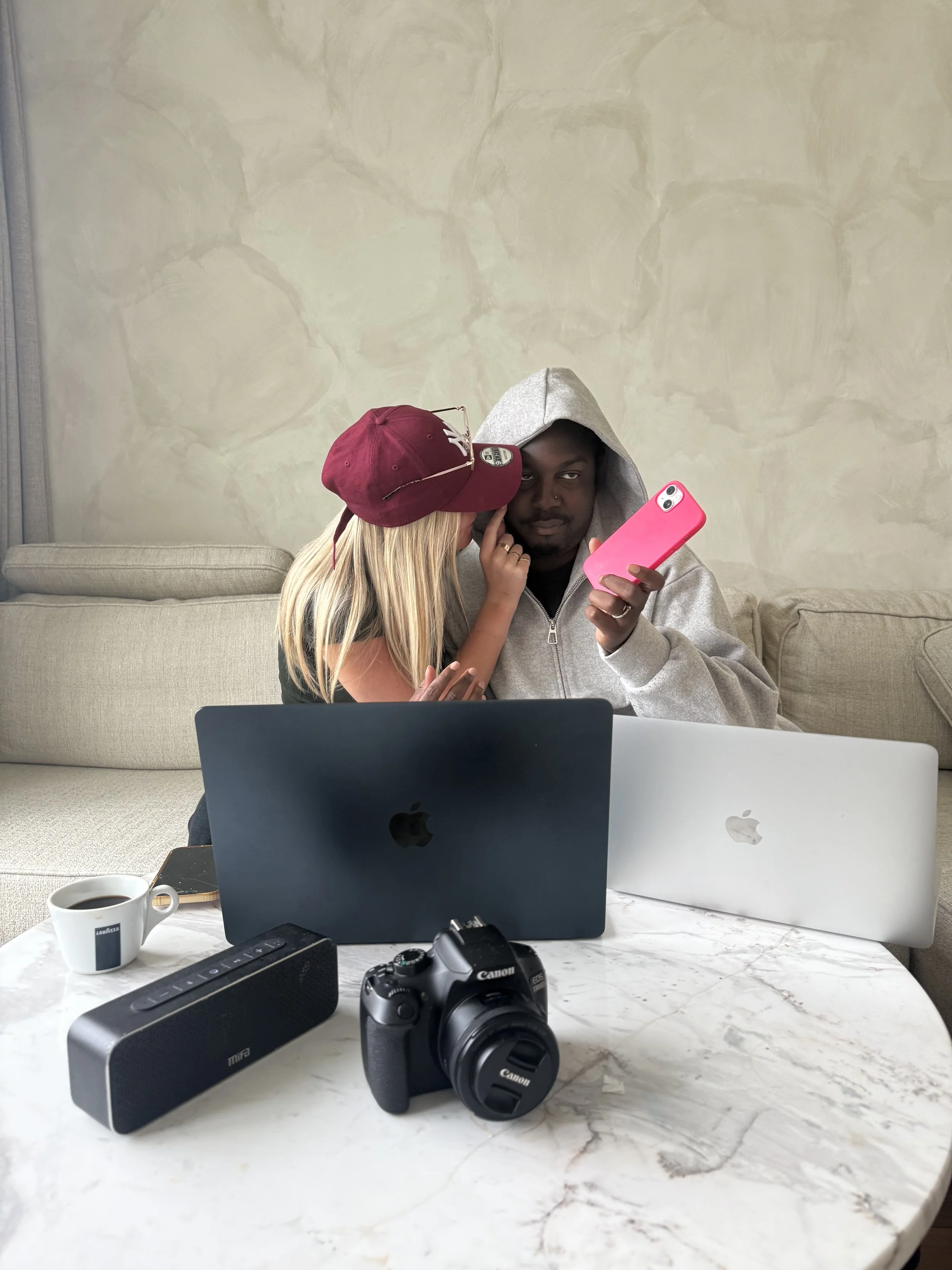 Two people, one with blonde hair wearing a burgundy baseball cap and the other with dark skin wearing a gray hoodie, are sitting close together, with the person in the hoodie looking at a pink smartphone held by the person in the cap. The setting appears to be indoors on a light-colored sofa, with a beige textured wall in the background. On the table in front, there are two laptop computers, a white coffee mug filled with coffee, a black portable speaker, a Canon camera, and a remote control.
