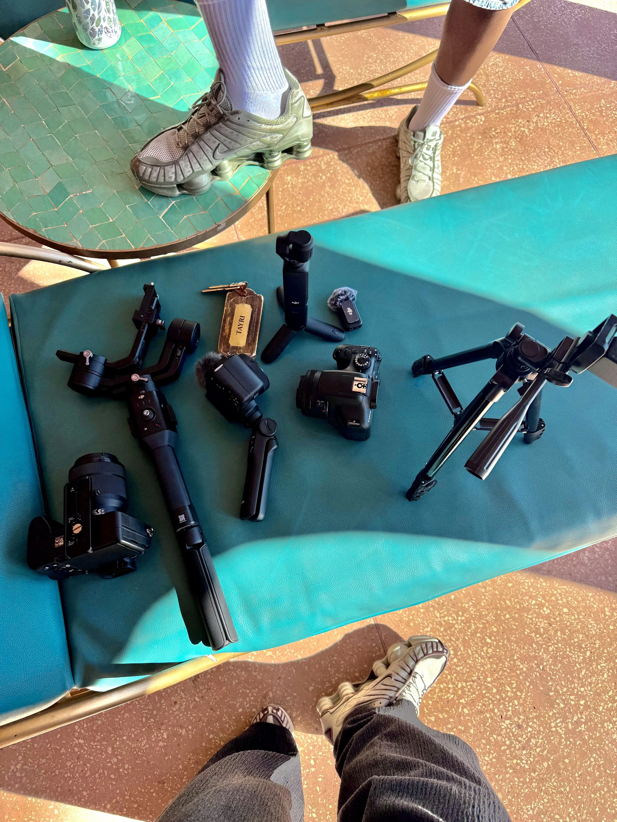 View of a person standing on the ground wearing sneakers and camouflage pants, with a teal bench in front of them. On the bench, there are various camera equipment, including cameras, a tripod, and other accessories. A teal cushion is placed on the bench, and a turquoise tiled surface is visible above the equipment. The ground is reddish-brown with small white specks, and the person's shadow is visible.