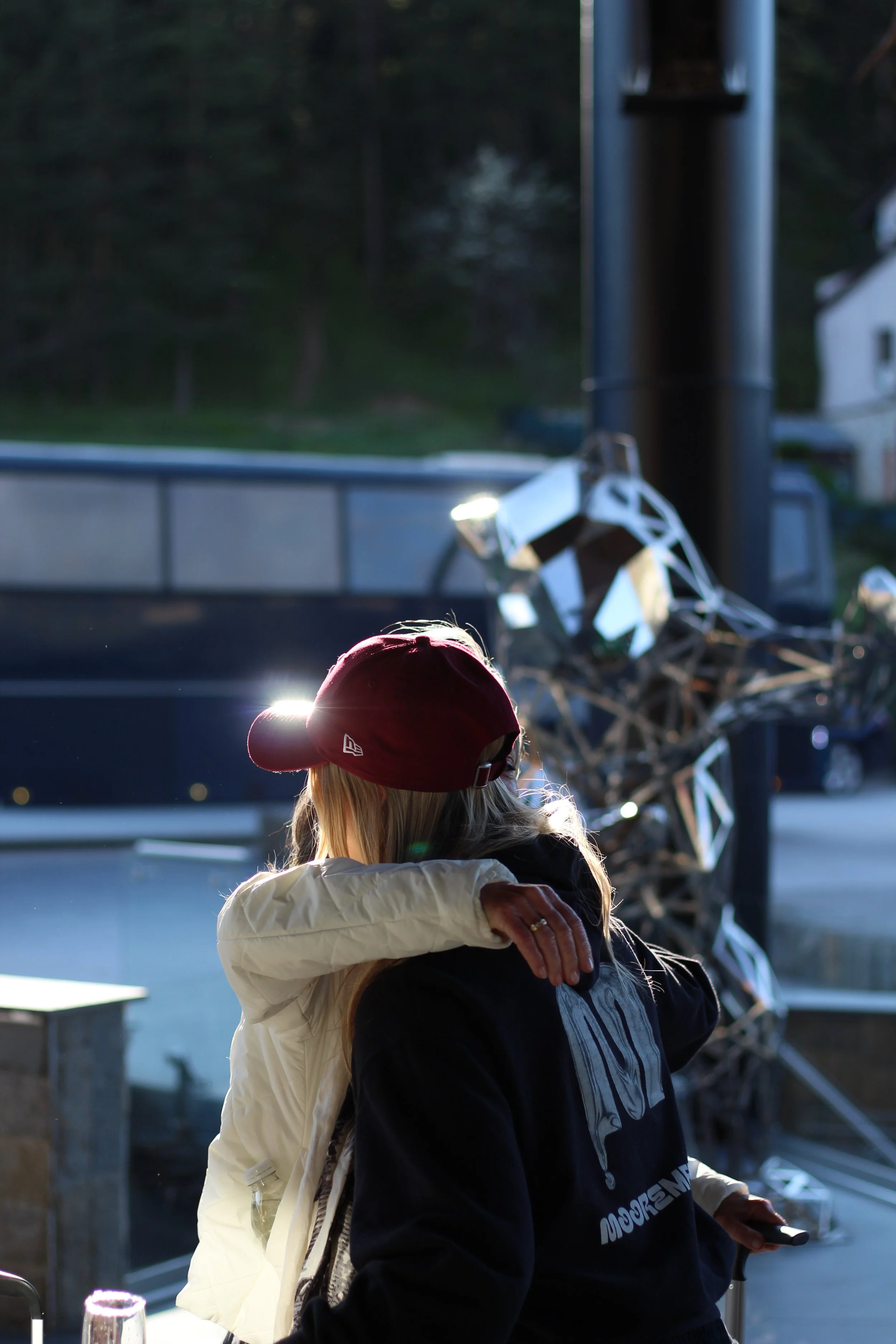 Two women hugging each other, one wearing a white jacket and maroon cap, the other in a black jacket with white lettering, with a vehicle and trees in the background during daytime.