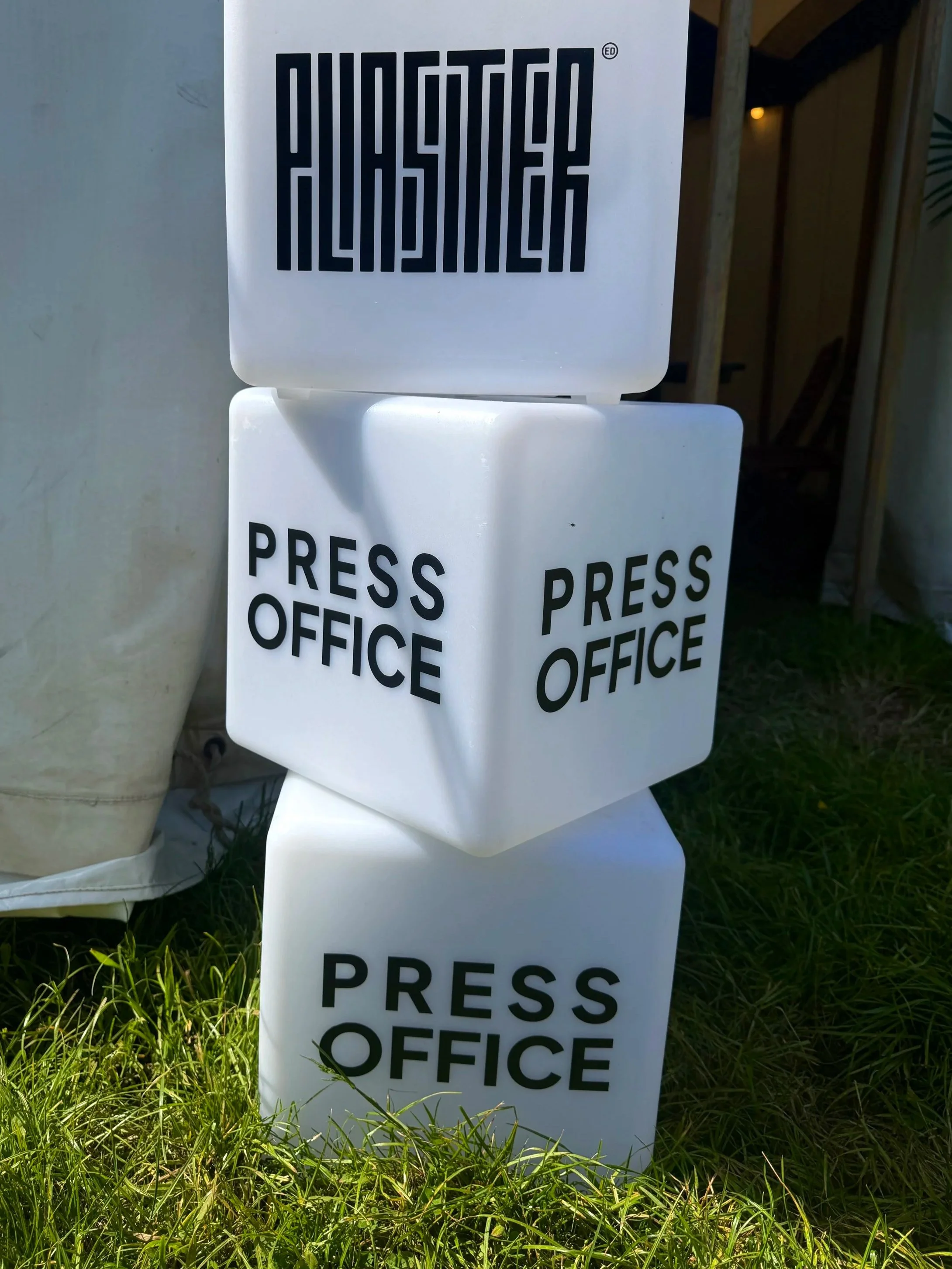 Multiple white signs with black text reading 'PRESS OFFICE' stacked on top of each other outdoors on grass, with part of a person and a wooden structure partially visible in the background.
