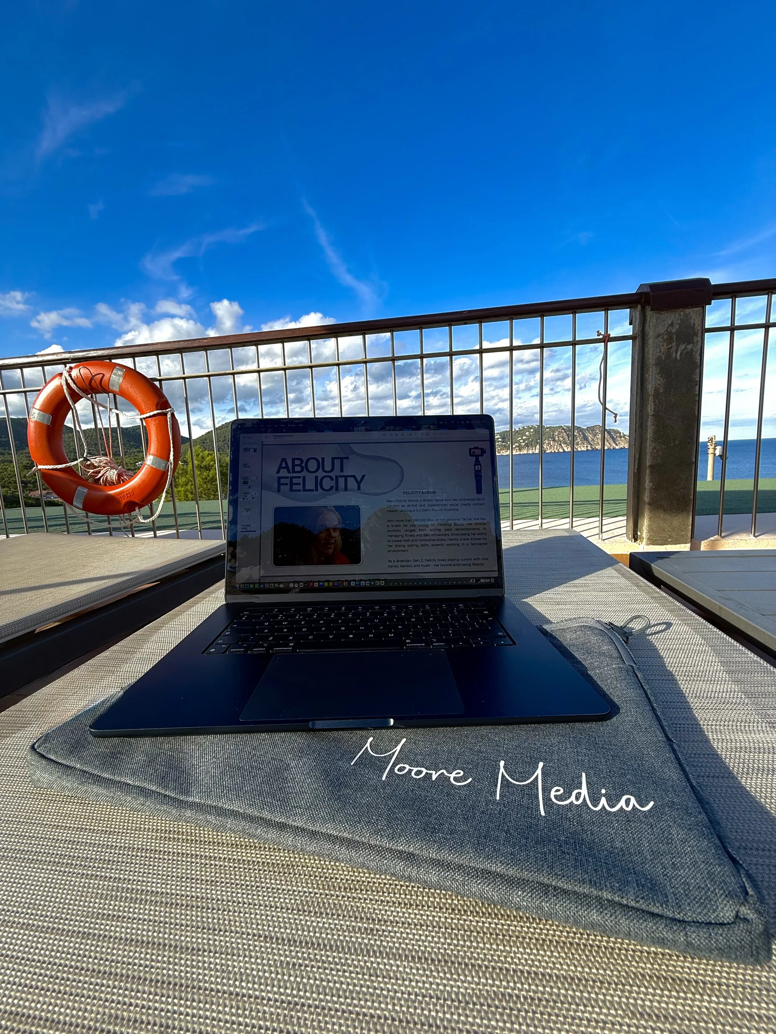 Laptop on outdoor table with view of blue sky, clouds, ocean, and hills in the background, with orange lifebuoy hanging on fence and a book or magazine open on the table.