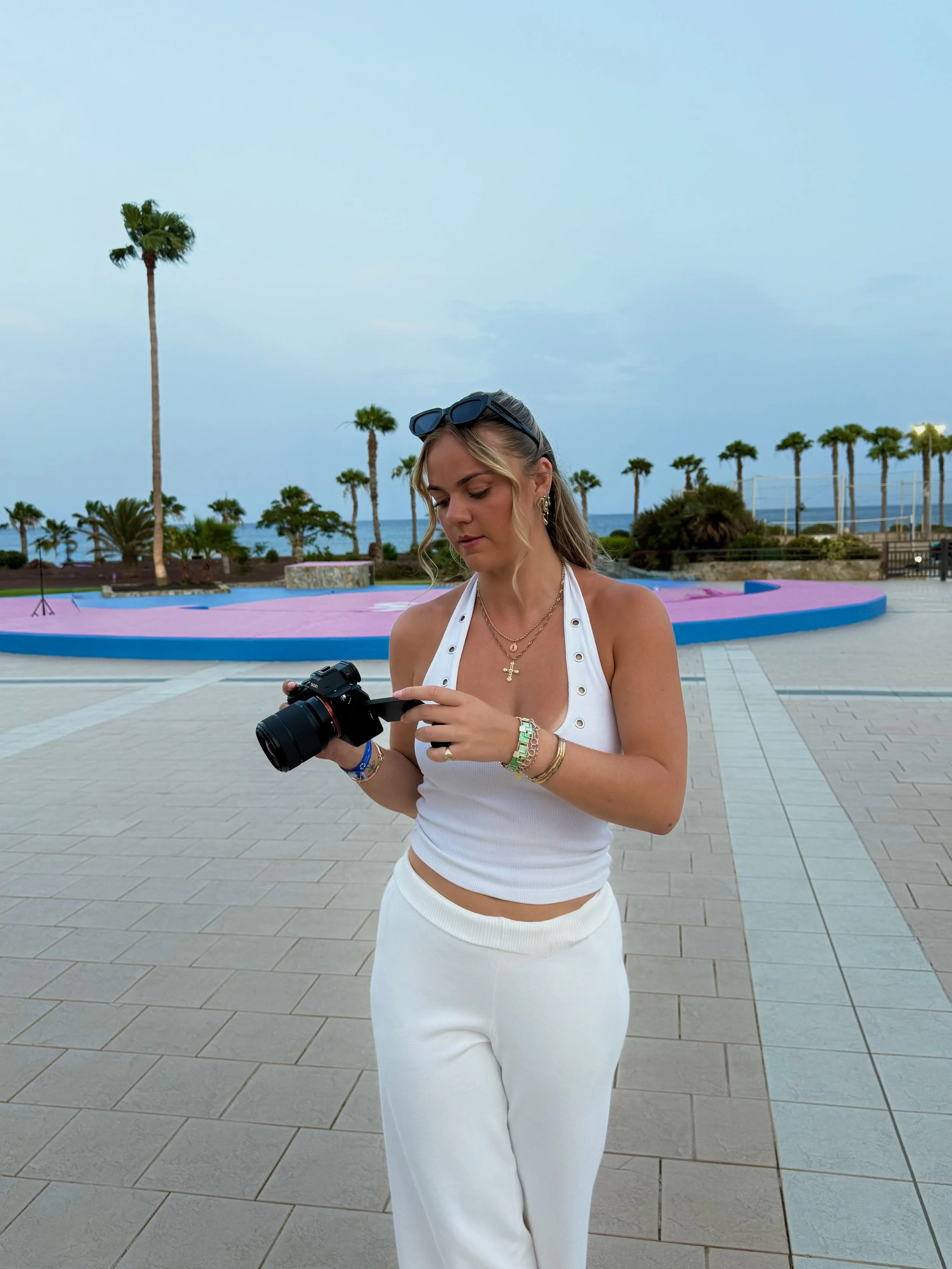 A woman in white pants and a white sleeveless top with sunglasses on her head is looking at a camera she is holding, standing in an outdoor area with pink and blue concrete surfaces, palm trees, and a view of the ocean in the background during the daytime.