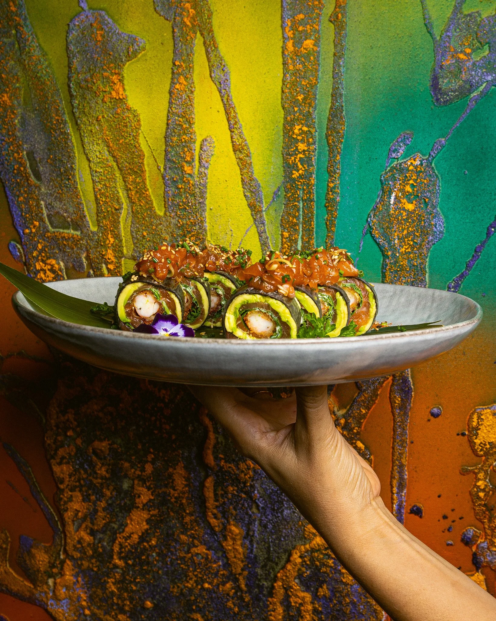 Prato de sushi roll com molho e sementes, sendo segurado por uma mão, com fundo de tecido decorado com cores vibrantes e padrões abstratos