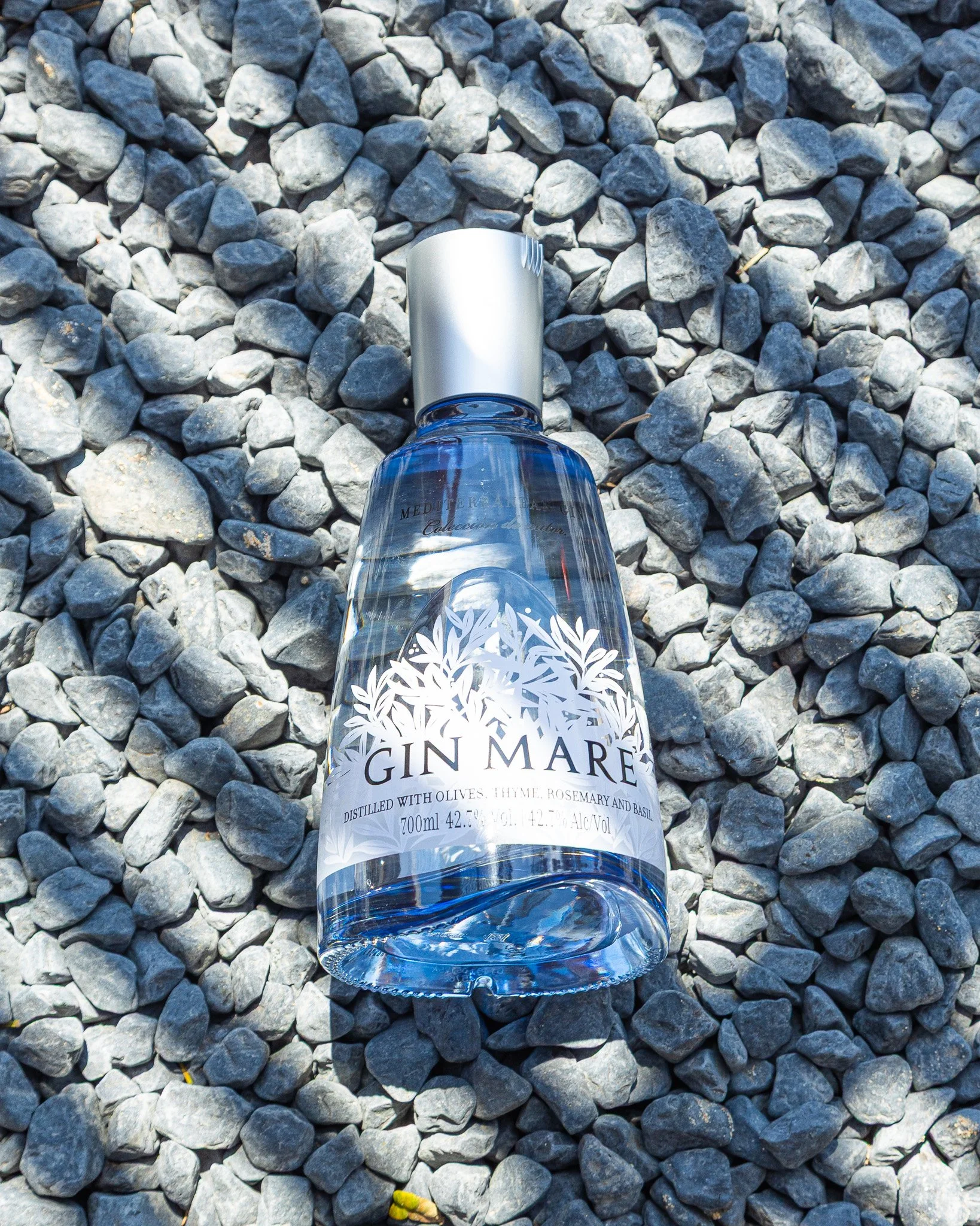 Garrafa de gin transparente com rótulo decorativo de folhas brancas, deitada sobre pedras cinzentas.