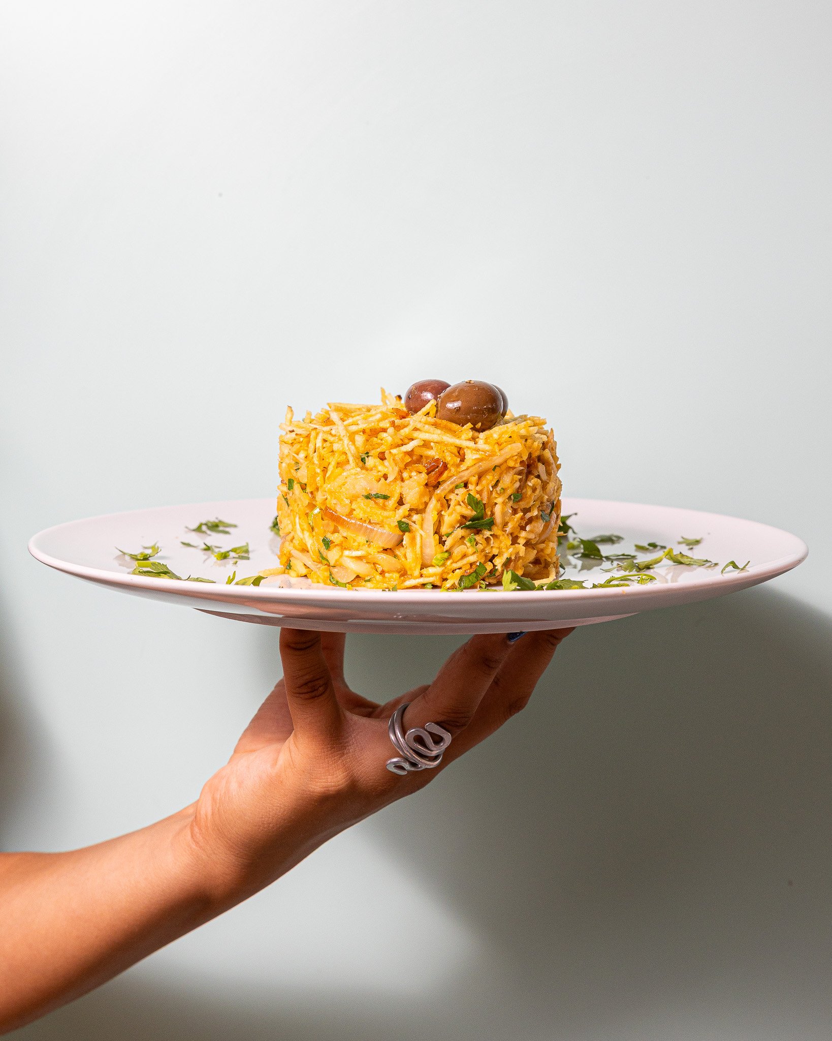 Pires branco com um prato de arroz com pedaços de cebola, decorado com folhas verdes, sendo segurado por mão feminina com anéis prateados.