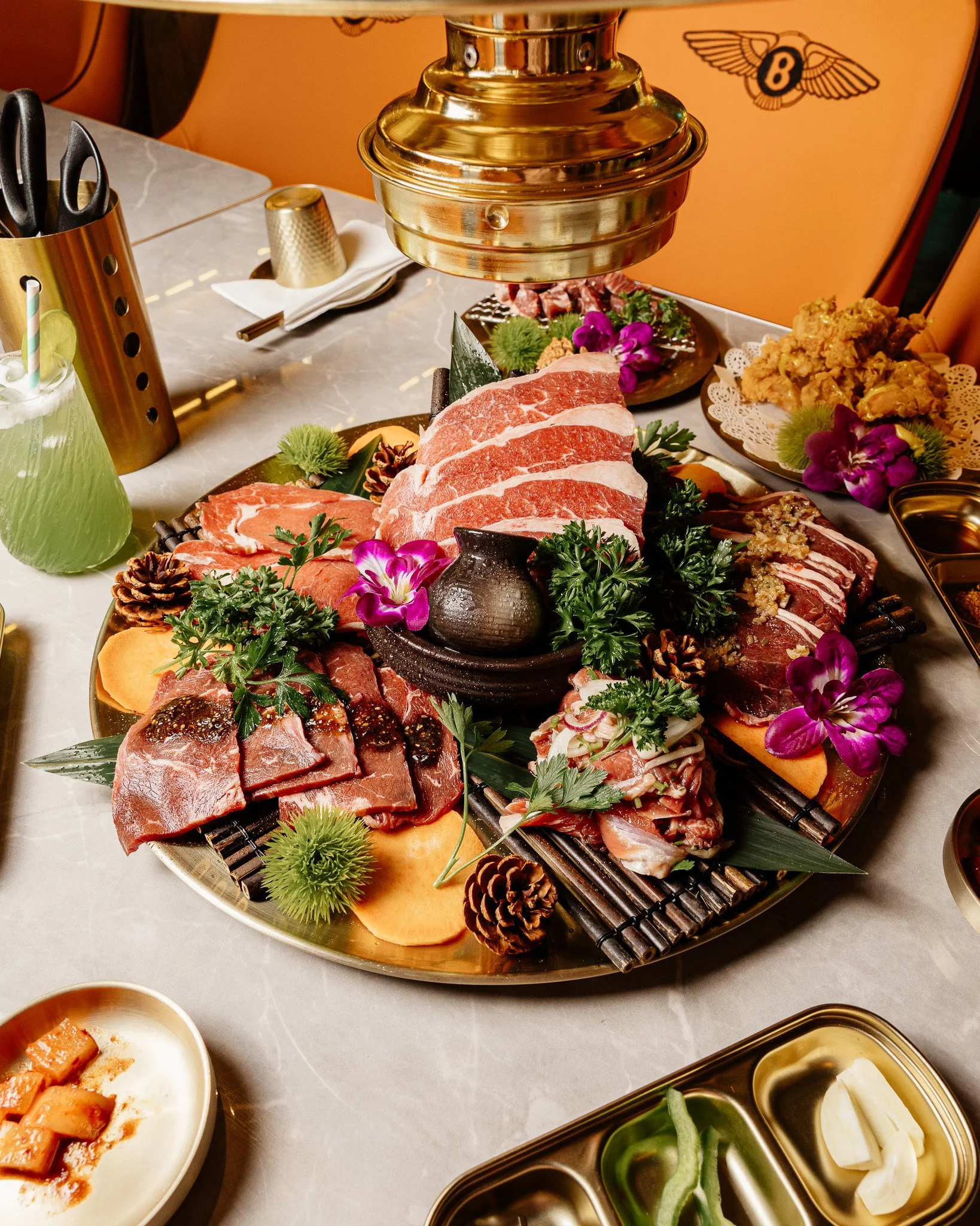 Prato de carnes variadas, incluindo cortes de carne vermelha, decorado com flores, folhas verdes, pinhas, e servido com acompanhamentos e aperitivos ao redor.