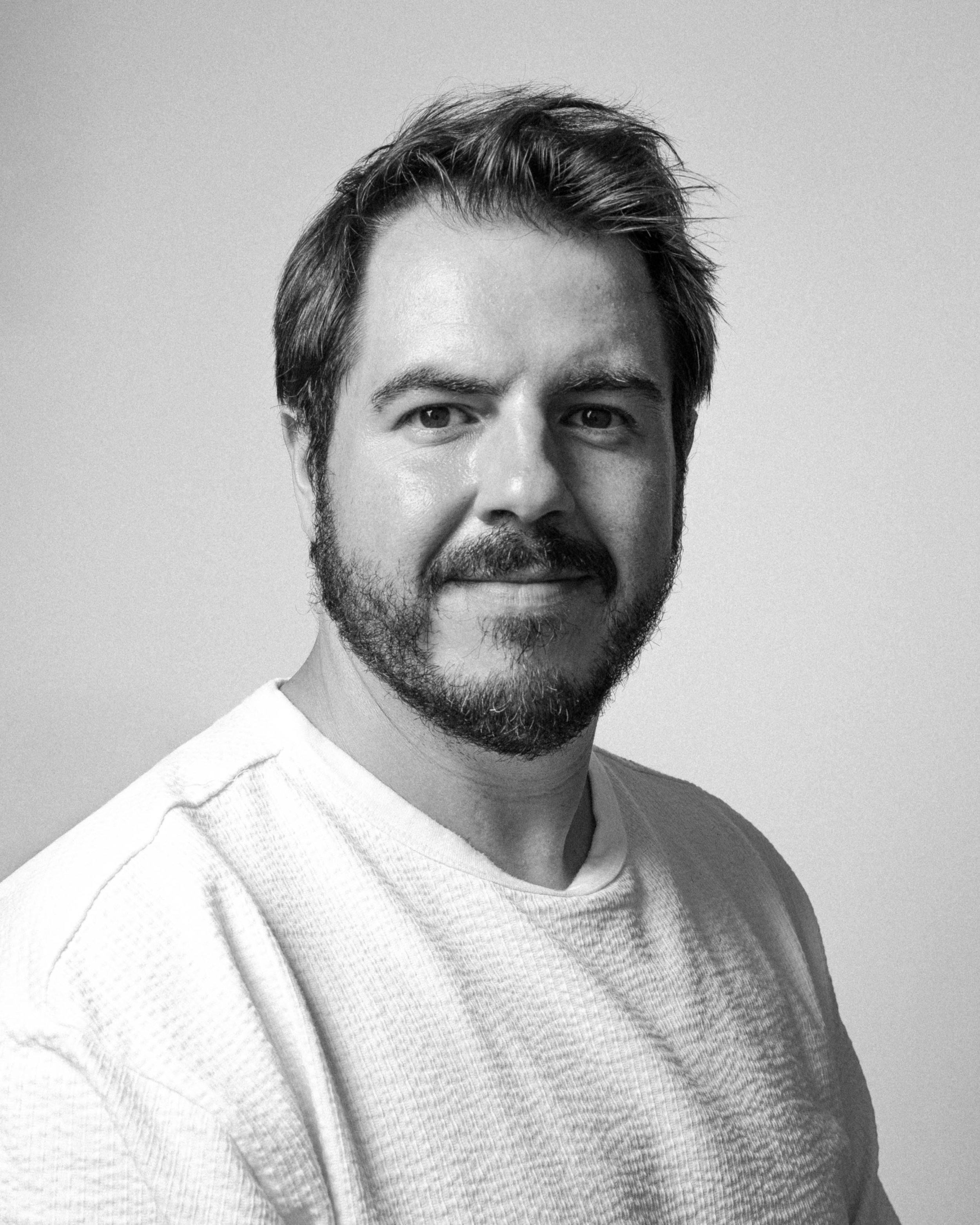 Retrato de um homem com cabelo e barba escuros, usando uma camiseta de malha cinza clara, em close-up. Foto em preto e branco.
