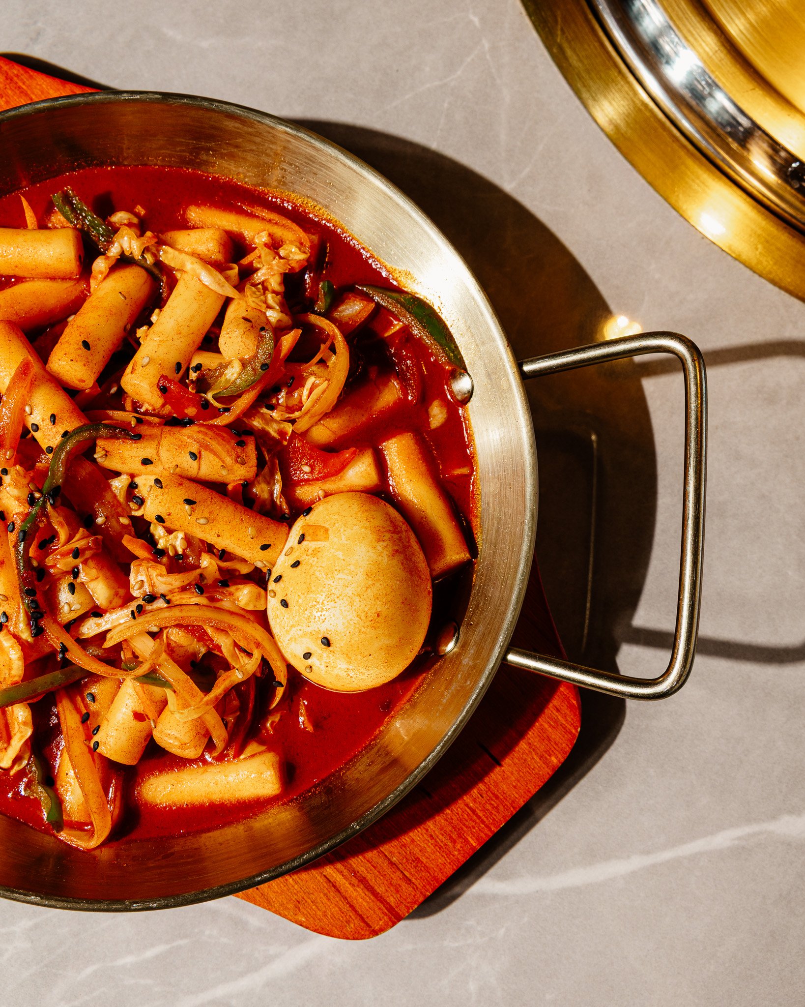 Panela de inox com comida coreana, incluindo rabokki com molho vermelho, ovos cozidos, bolinhos, cenoura, couve, manga, com sementes de gergelim preto.