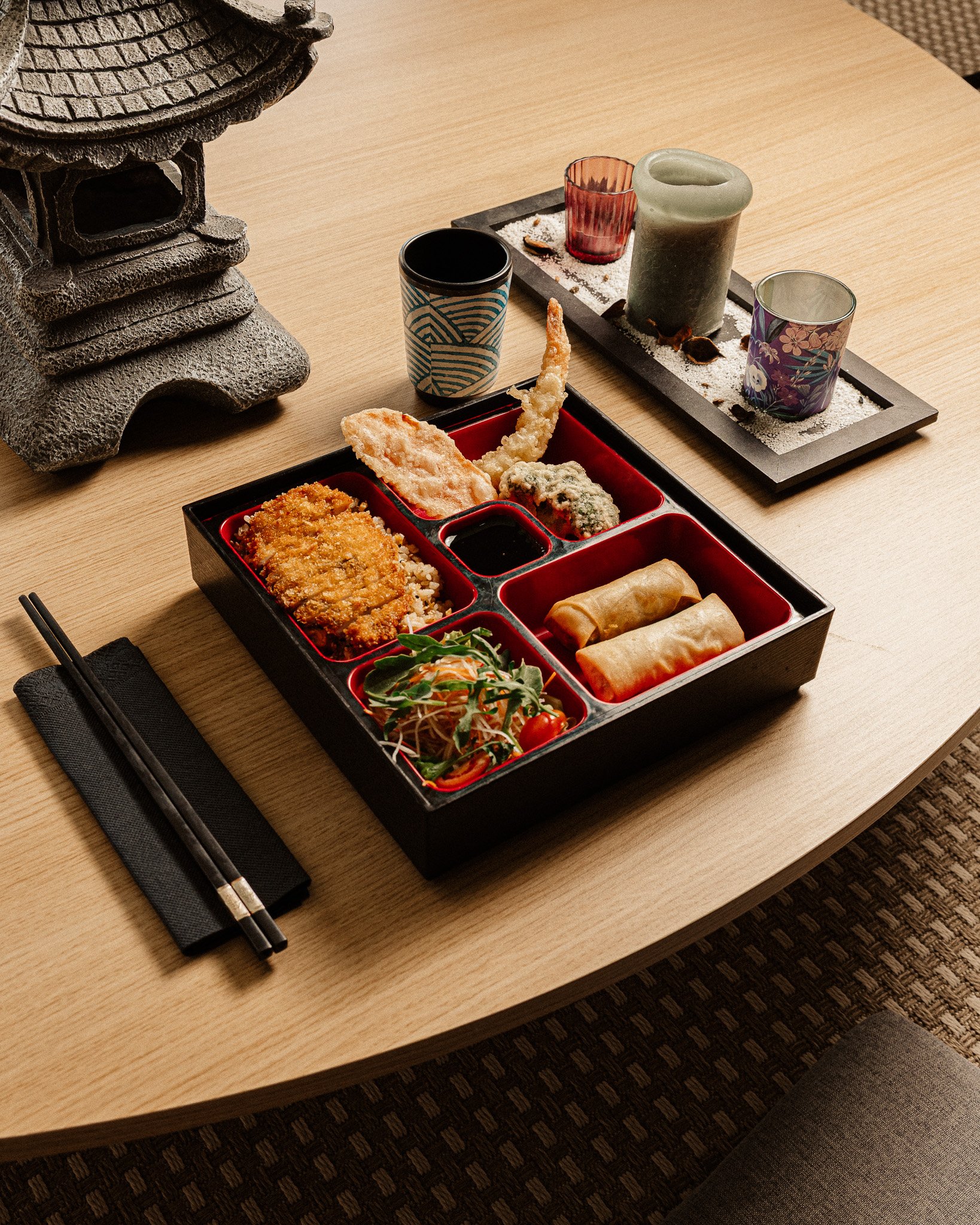 Mesa de madeira com um bento tradicional de comida japonesa, contendo diferentes pratos, além de utensílios de palito e um prato de salada. Ao fundo, uma decoração com velas, vasos e areia branca.