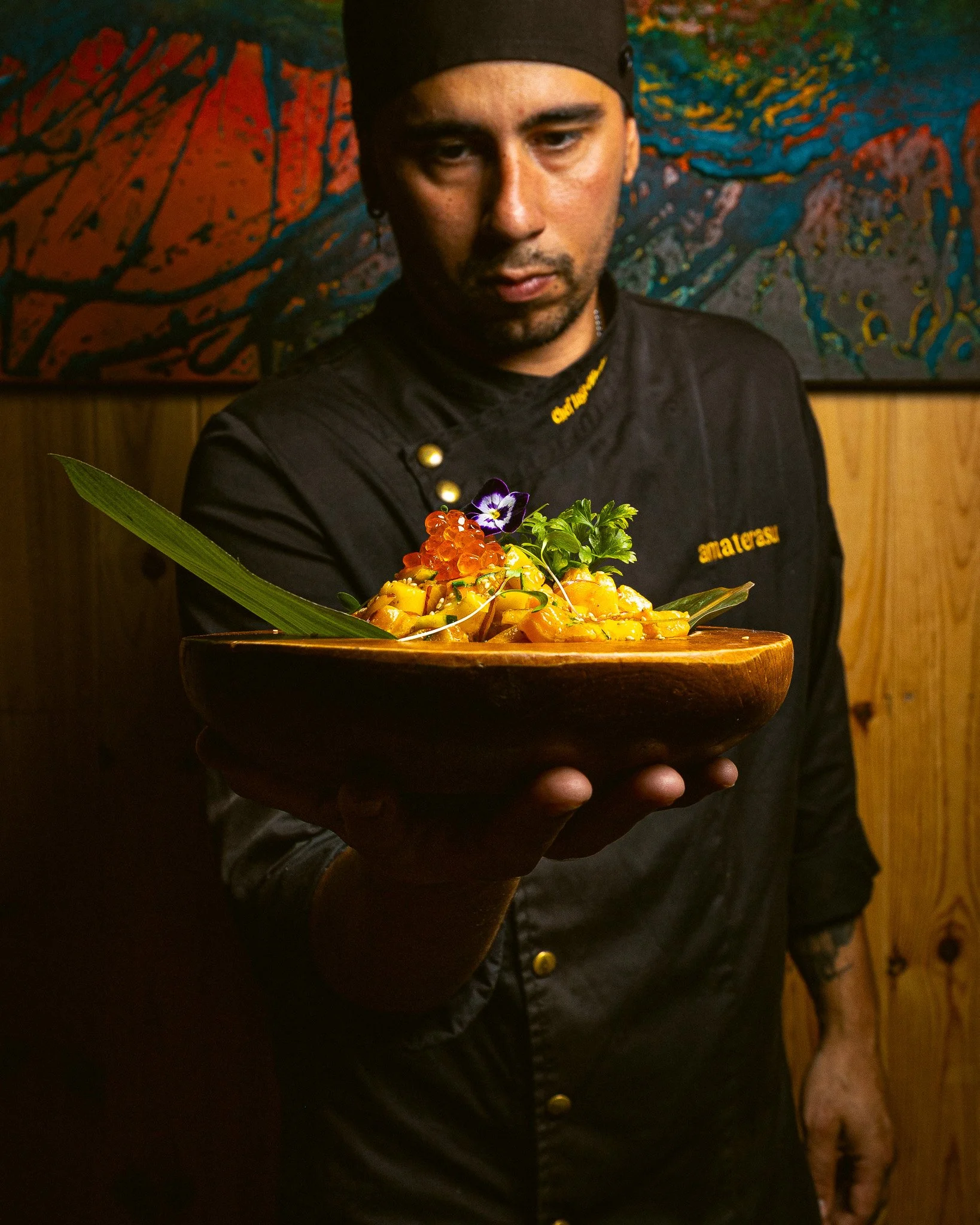Homem apresentando um prato de comida artesanal decorado com flores, usando avental preto com detalhes em dourado, em um ambiente com fundo colorido e parede de madeira.