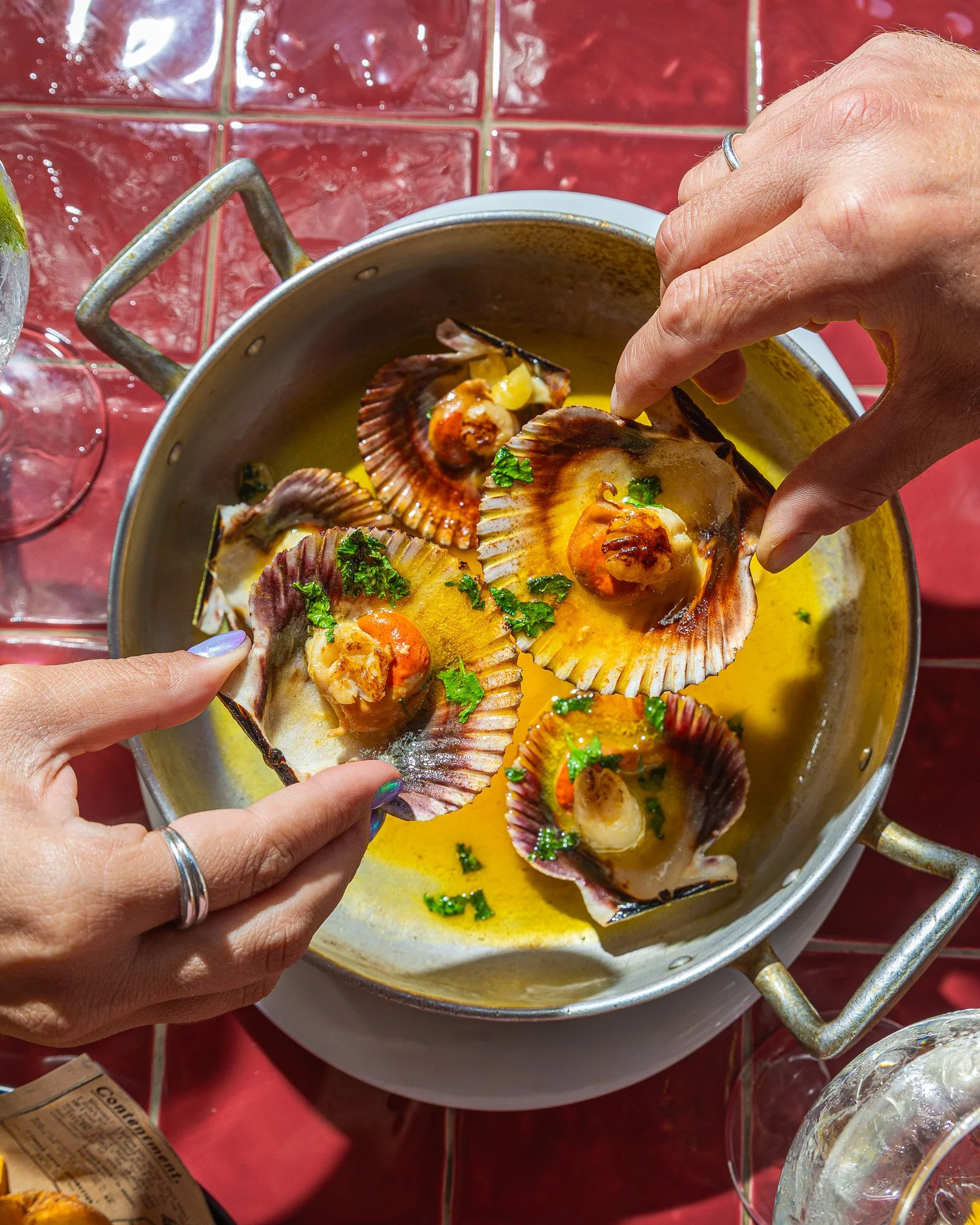 Pessoa segurando casquinha de vieira recheada com molho, cebola e salsa, sendo colocada em uma panela de fondue de queijo. Ao fundo, utensílios de cozinha e uma mesa com toalha vermelha.