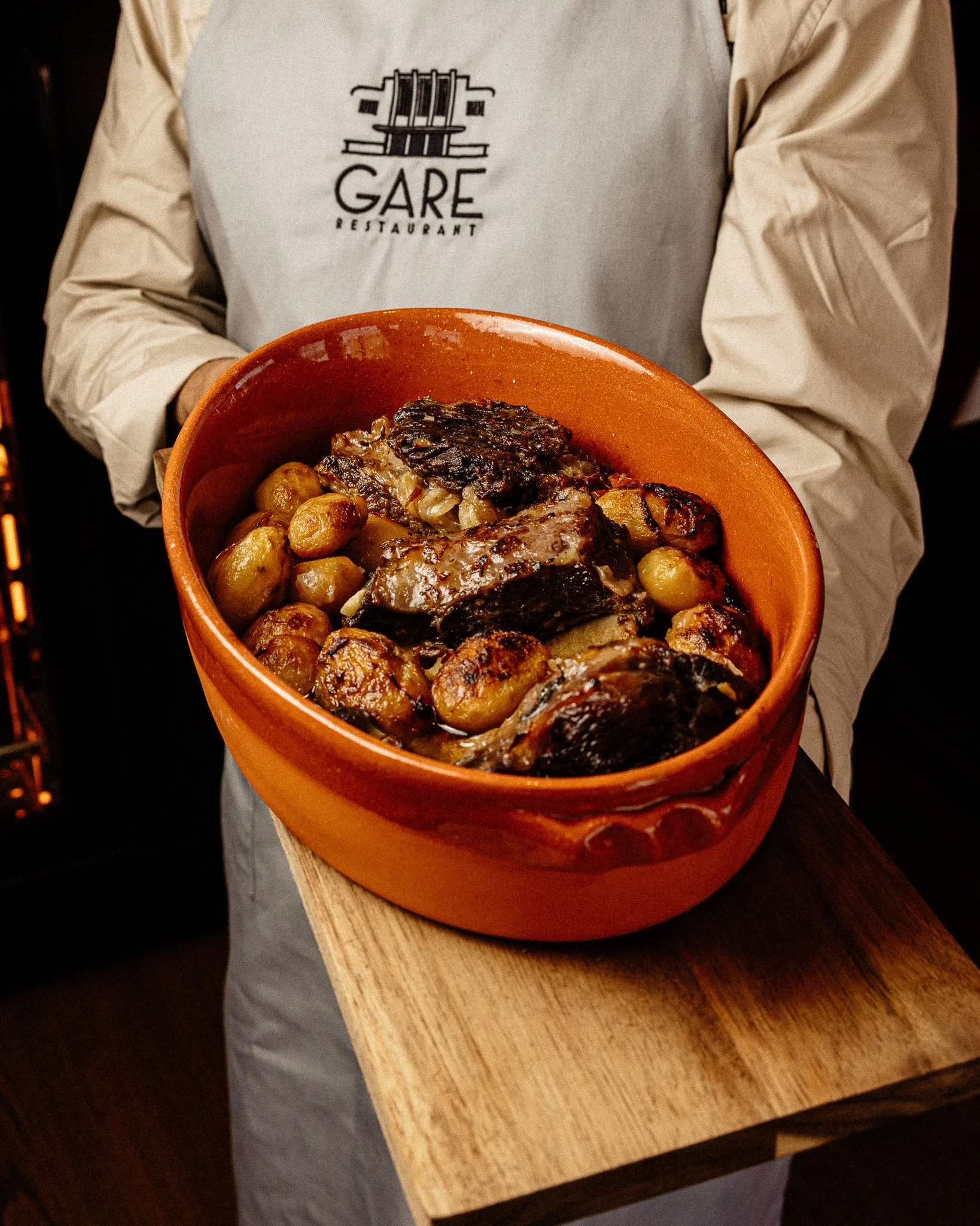 Pessoa segurando uma tigela de cerâmica com um ensopado de carne, batatas e legumes. A pessoa usa um avental que tem o logo do restaurante Gare.