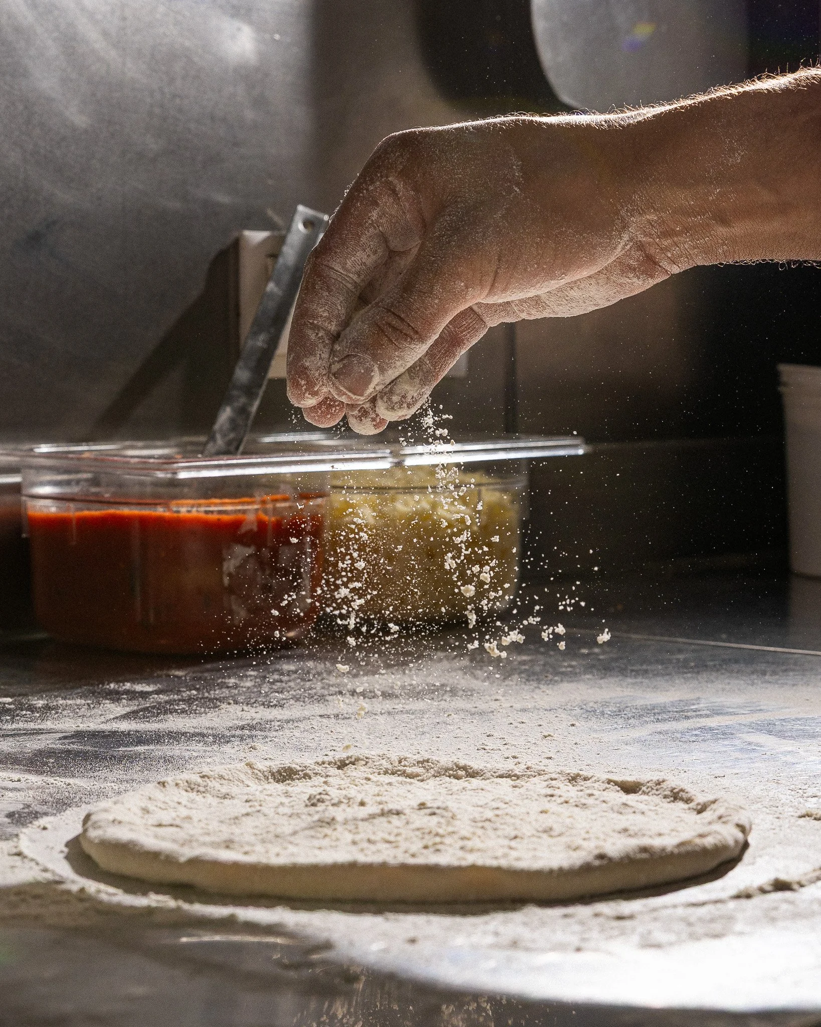Imagem de uma mão polvilhando farinha sobre uma massa de pizza crua em uma superfície de cozinha, com uma forma de pizza recortada ao lado.