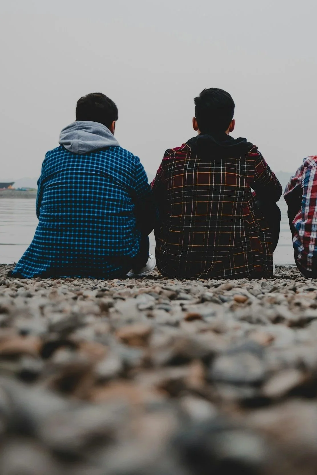 Three young men sitting on a rocky beach facing a body of water on a cloudy day, with their backs to the camera.