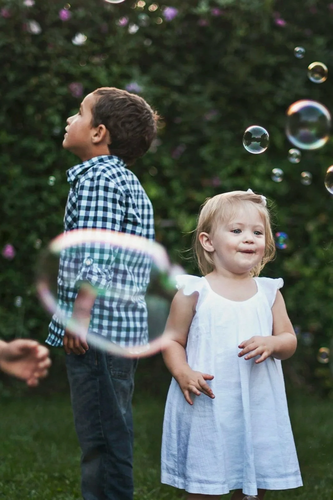 Two young children, a boy and a girl, playing outdoors among floating soap bubbles. The boy is wearing a blue checkered shirt and dark pants, looking up with a curious expression. The girl is dressed in a white dress, looking slightly to the side with a playful expression. Green foliage is in the background.